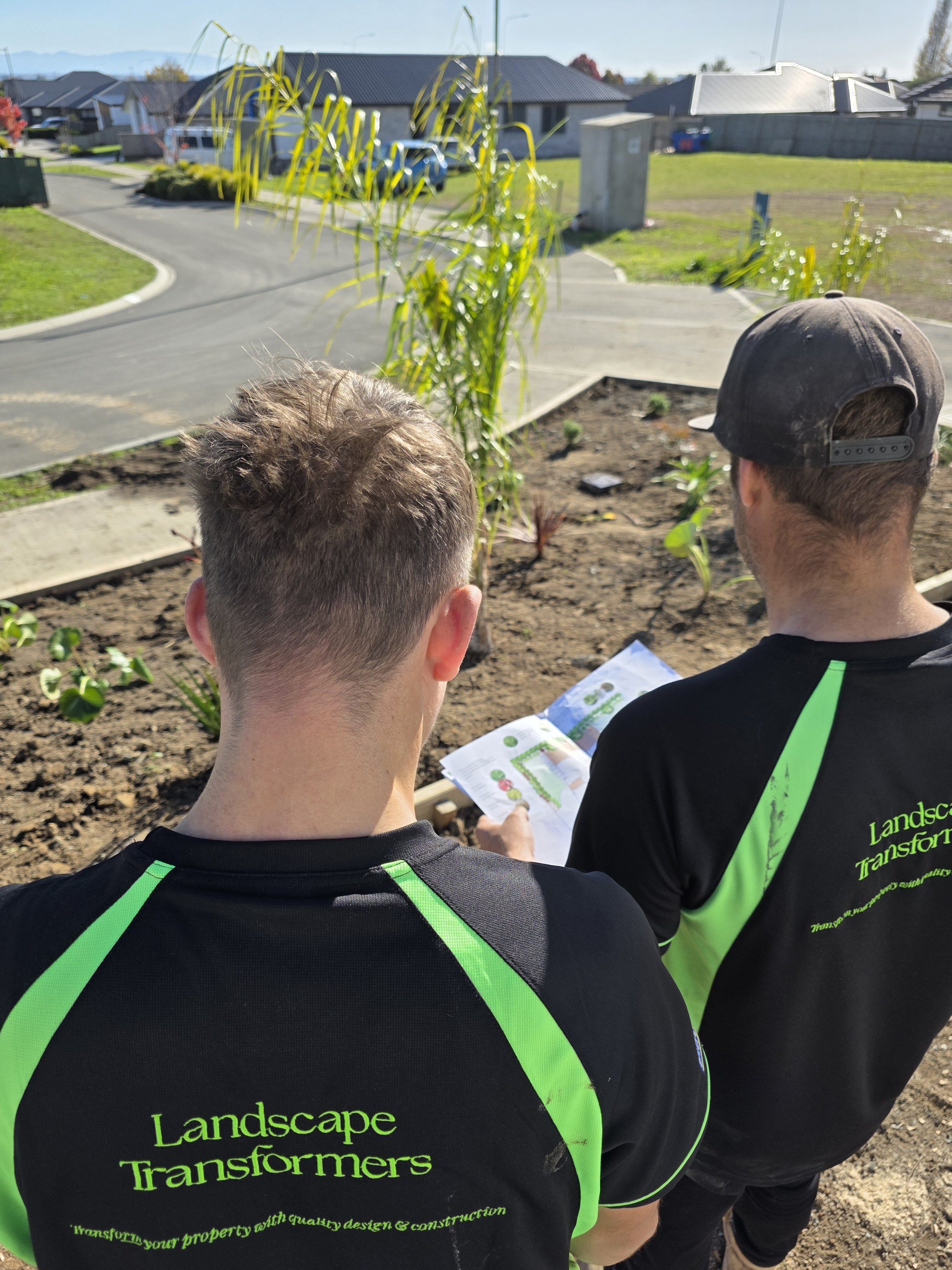 Two men wearing black T-shirts with green accents and the text 'Landscape Transformers' on the back are standing outdoors on a garden bed, looking at a map or plan. Behind them, there are small plants, a paved driveway, and residential houses under a