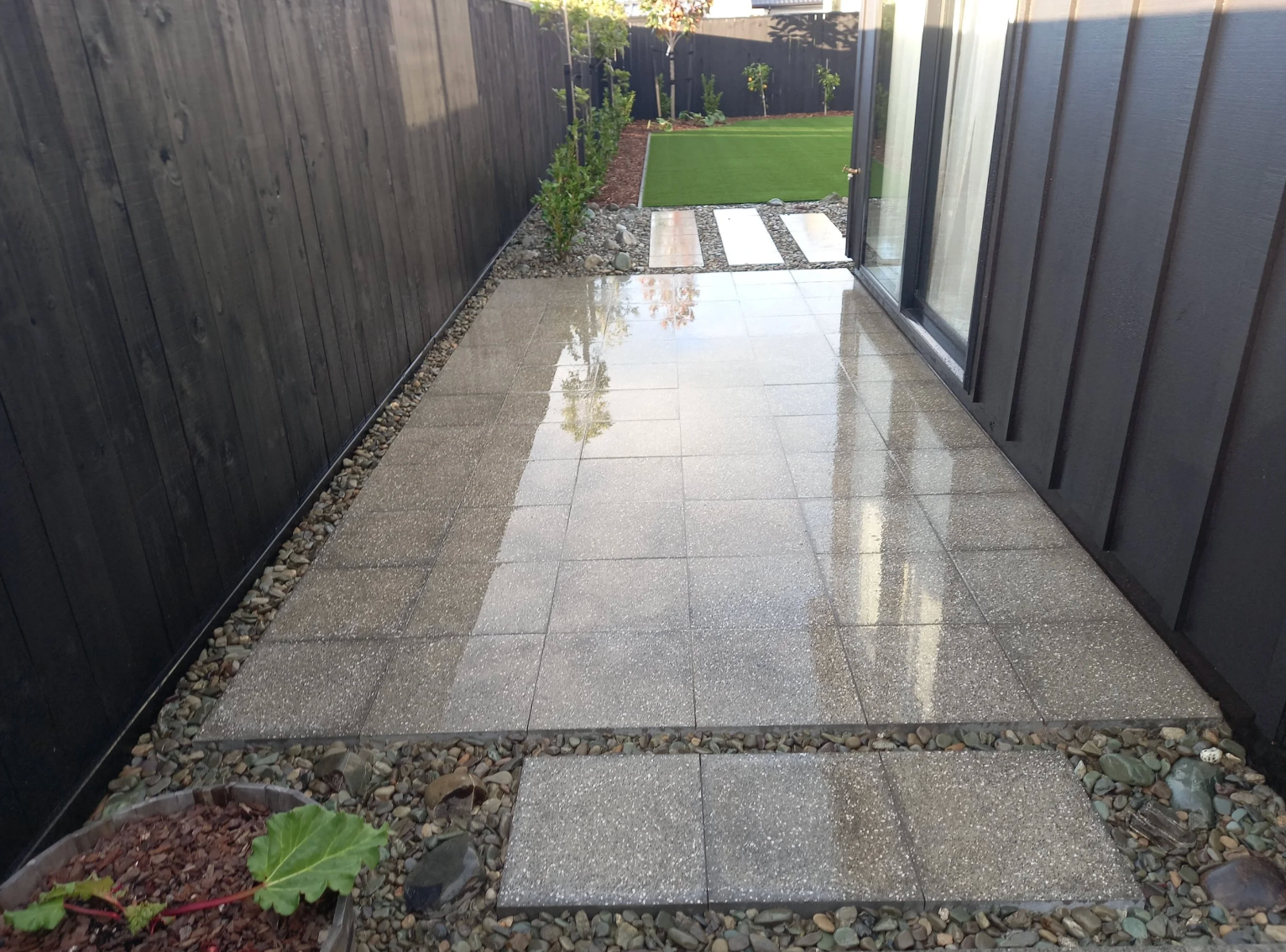 Rain-soaked patio with large tiled area leading to a grassy backyard with small plants and trees, enclosed by black fencing.
