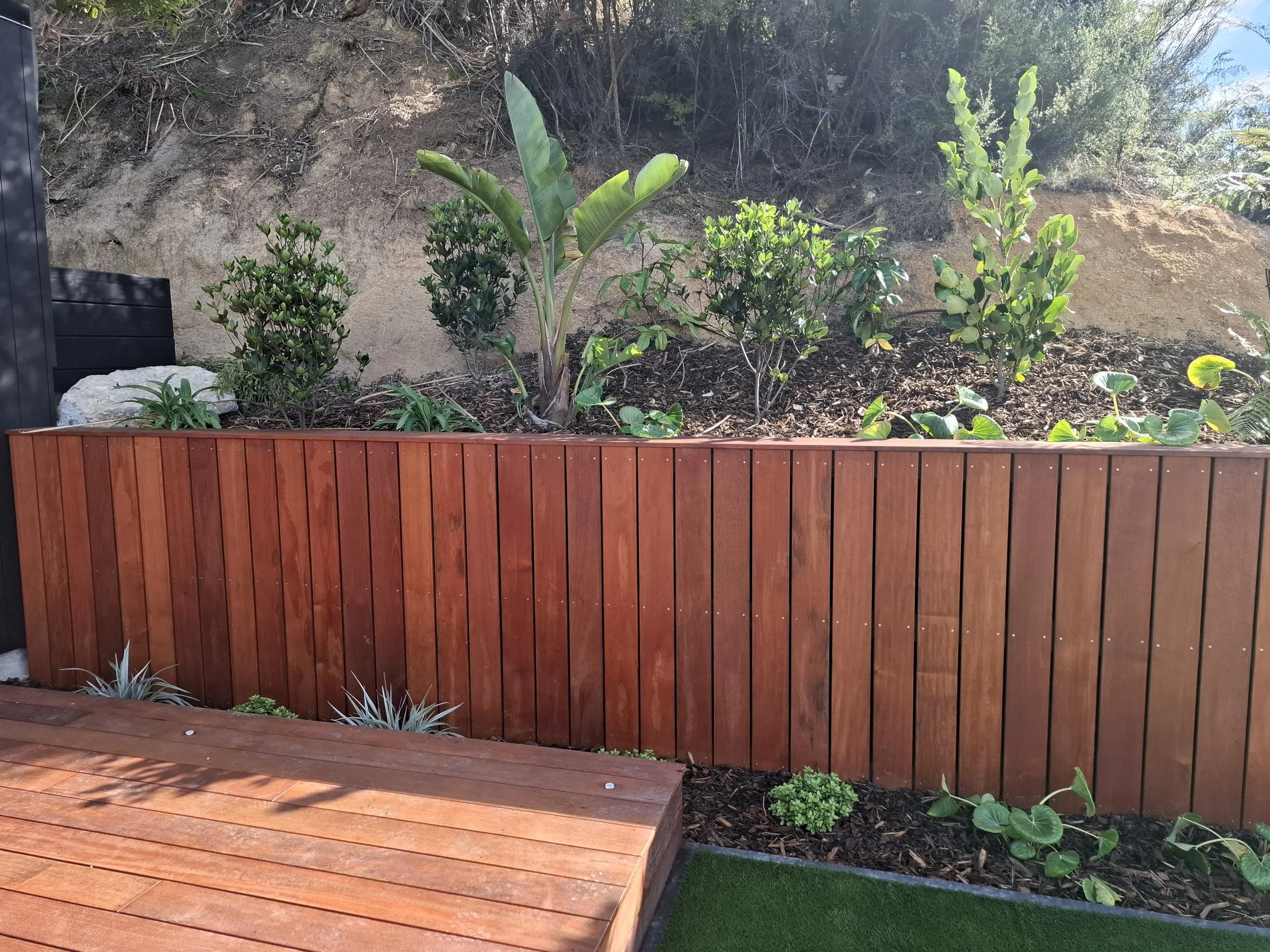 A garden with a raised wooden bed, green plants, and a dirt hillside in the background. There is also artificial grass and a black planter box visible.