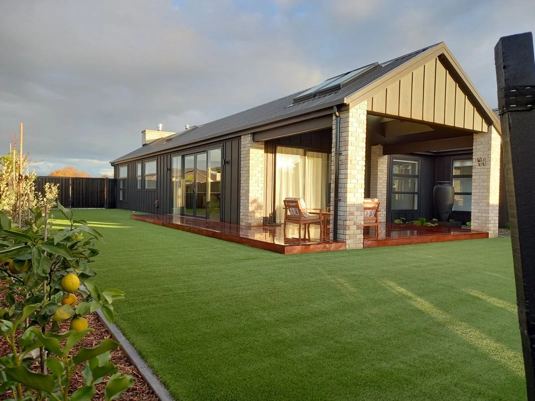 Modern house with a large backyard, wooden porch with chairs, brick and dark siding exterior, large sliding glass doors, and an outdoor urn decoration under a pitched roof.