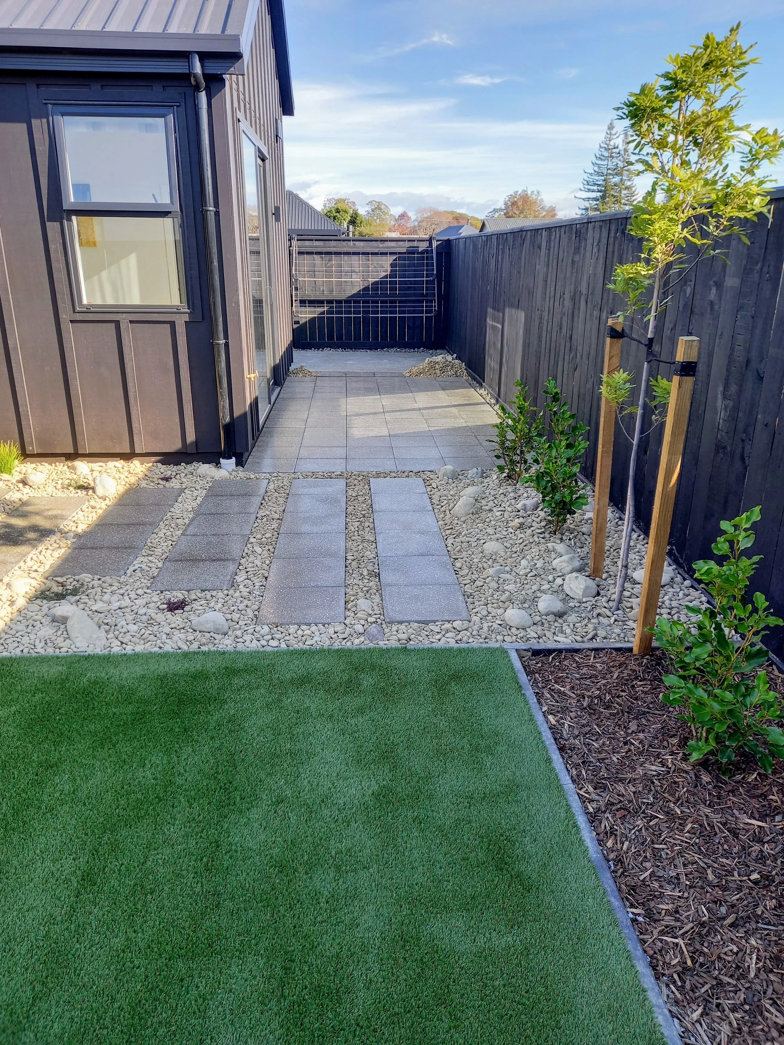 A backyard with a small paved patio area, surrounded by a black wooden fence, a young tree supported by stakes on the right, and a well-maintained grass lawn on the left. The sky is blue with some clouds, and neighboring houses are visible in the bac