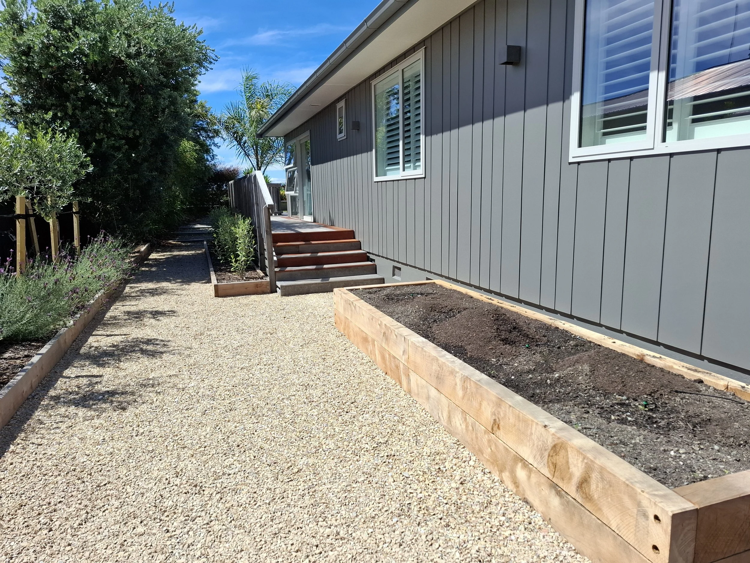 A modern house with gray vertical siding, white-framed windows with shutters, and a small wooden staircase leading to a sliding glass door. There is a gravel pathway along the house, bordered by planter boxes, with lush green trees and plants nearby 