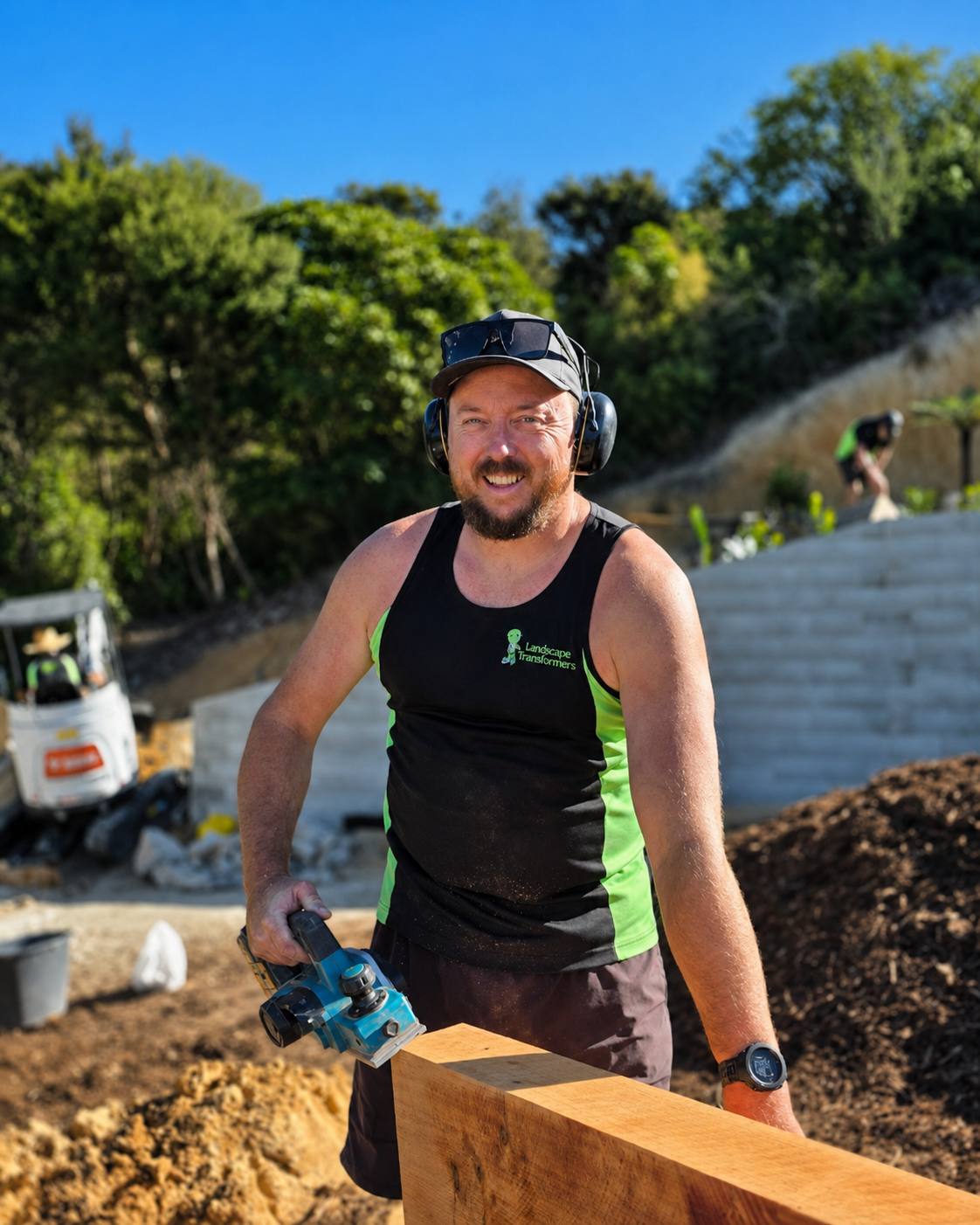 Man wearing safety headphones, sunglasses on a cap, and a black sleeveless shirt with green accents, holding a power saw, working outdoors on a construction site with dirt, wood, and construction equipment in the background.