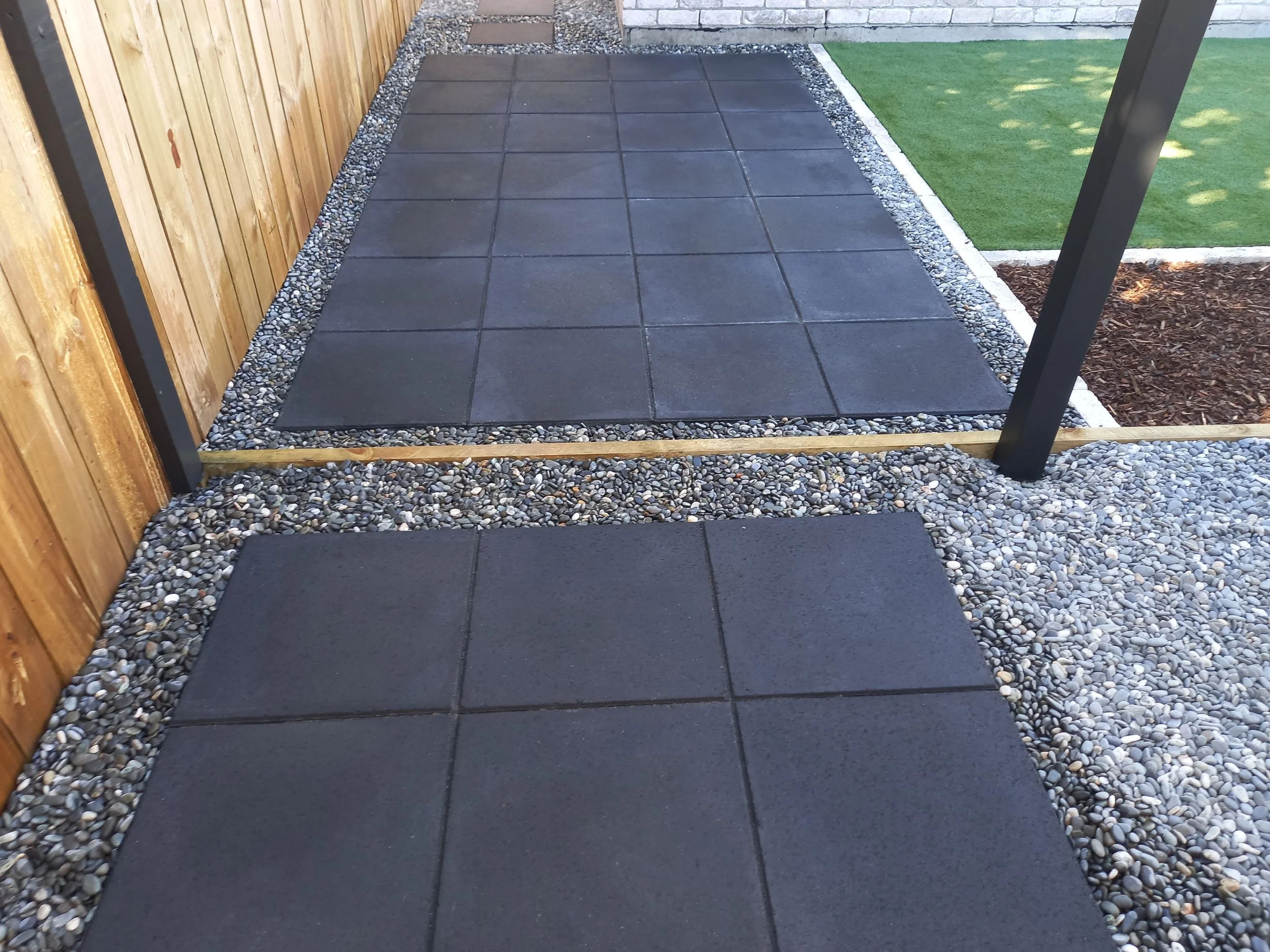 A newly landscaped backyard patio with large dark grey square tiles surrounded by small white gravel, with a wooden fence on the left and a grass lawn on the right.