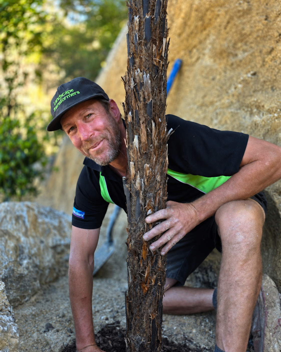 A man squatting outdoors, holding a young tree with a muddy trunk, wearing a black cap and a black shirt with green accents.