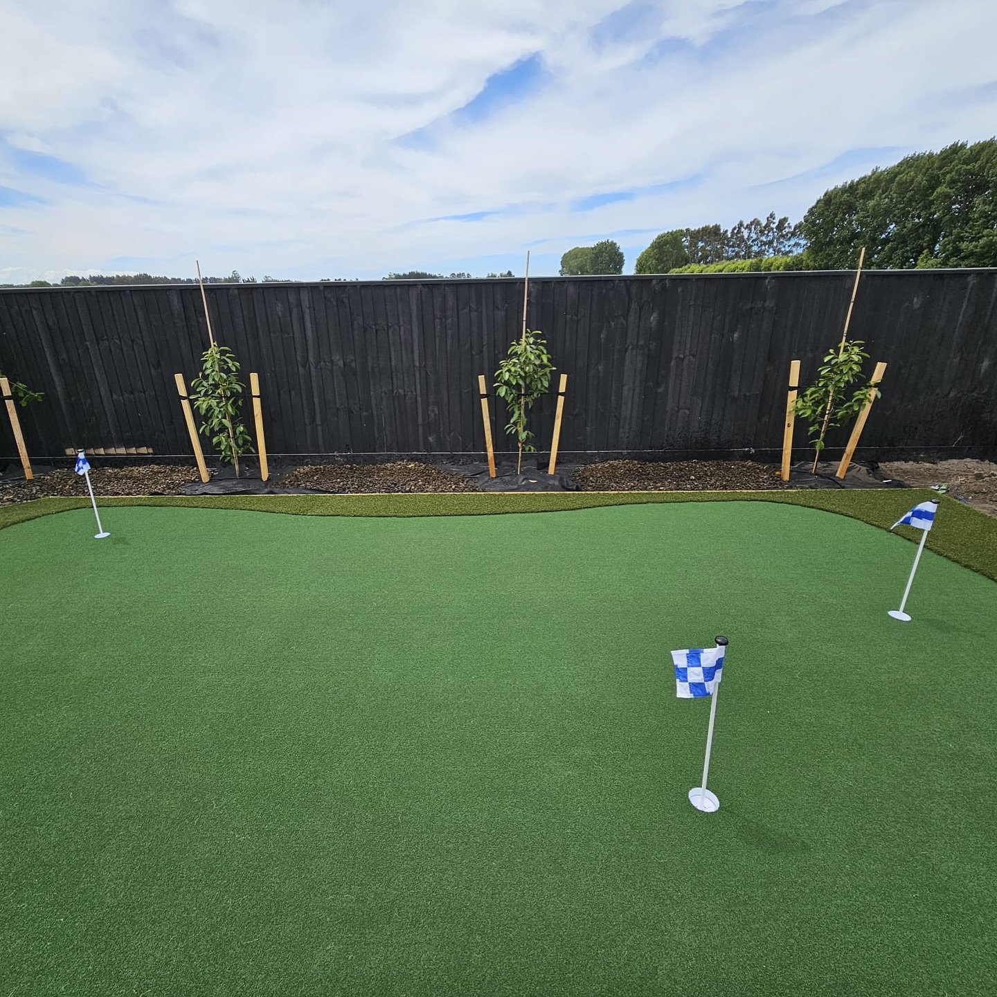 Miniature golf course with artificial turf, four flags marking holes, and young trees planted along a black wooden fence under a partly cloudy sky.