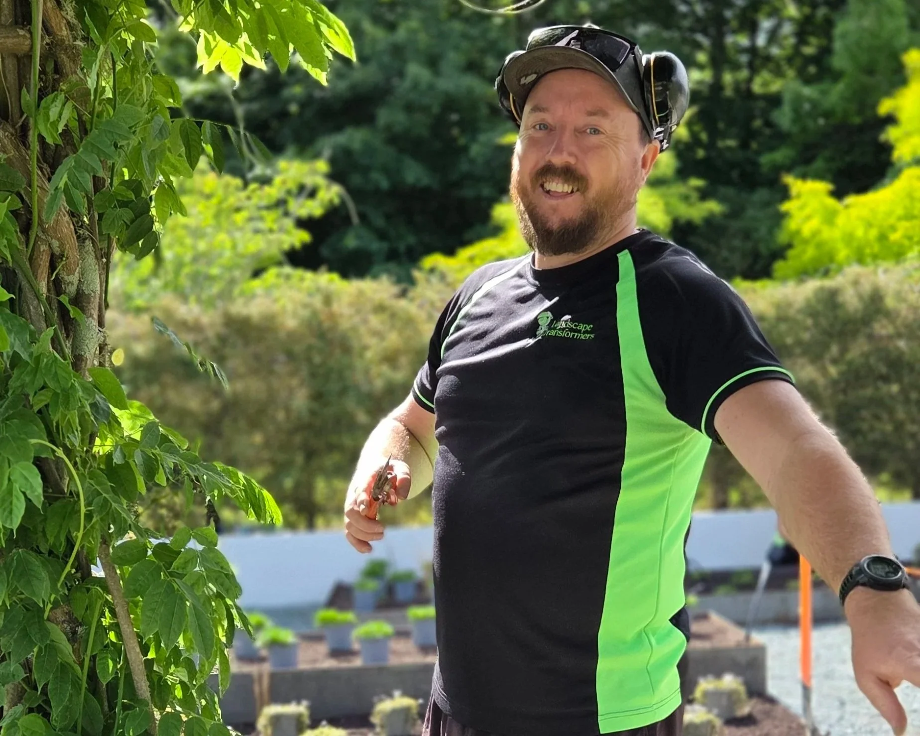 A man wearing a black and green T-shirt, a cap, and safety ear protection is smiling outdoors beside a leafy tree. He is holding pruning shears.