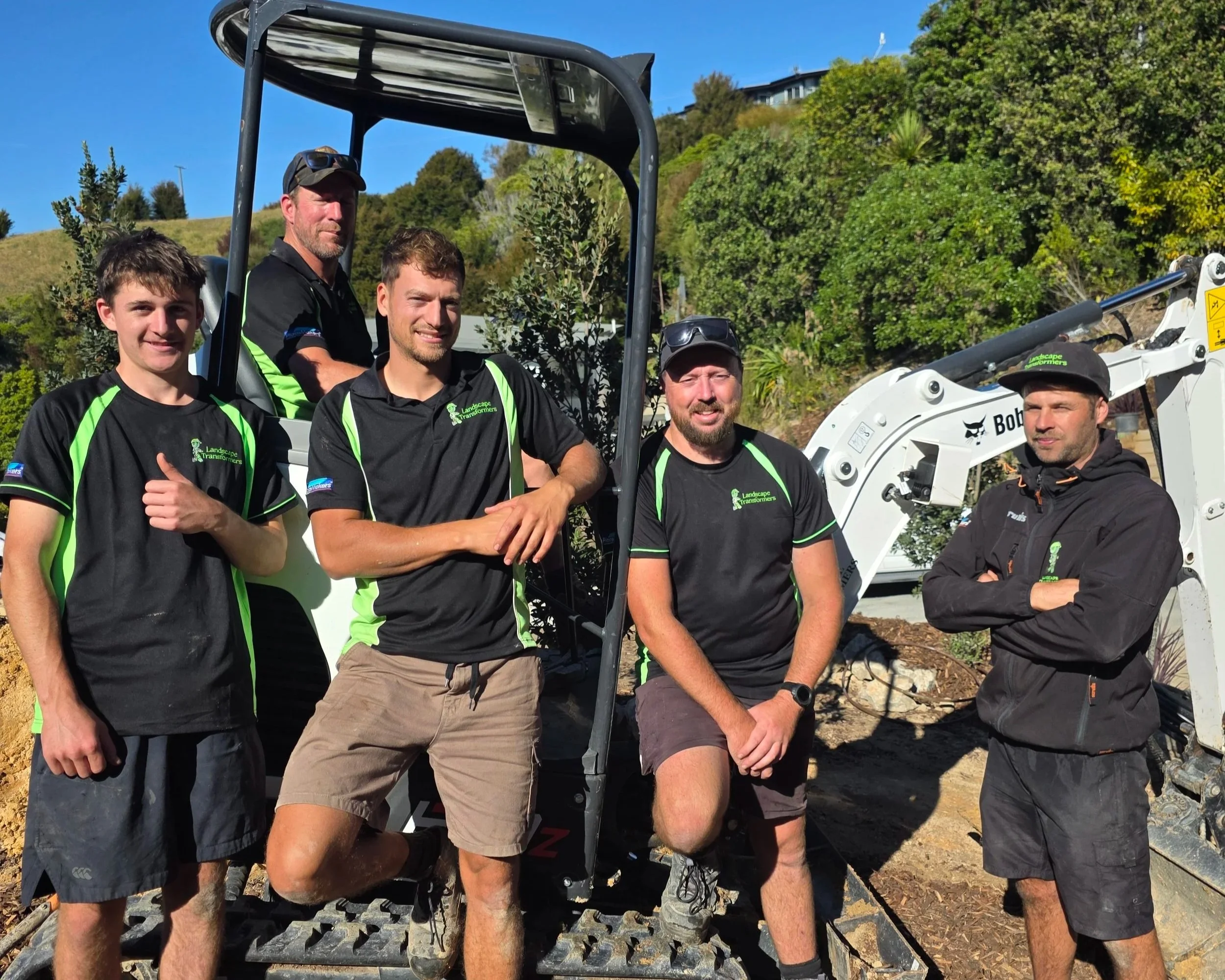 Group of five men standing outdoors near construction equipment, wearing black and green shirts with 'Landscape Transformers' logo, with greenery and a blue sky in the background.