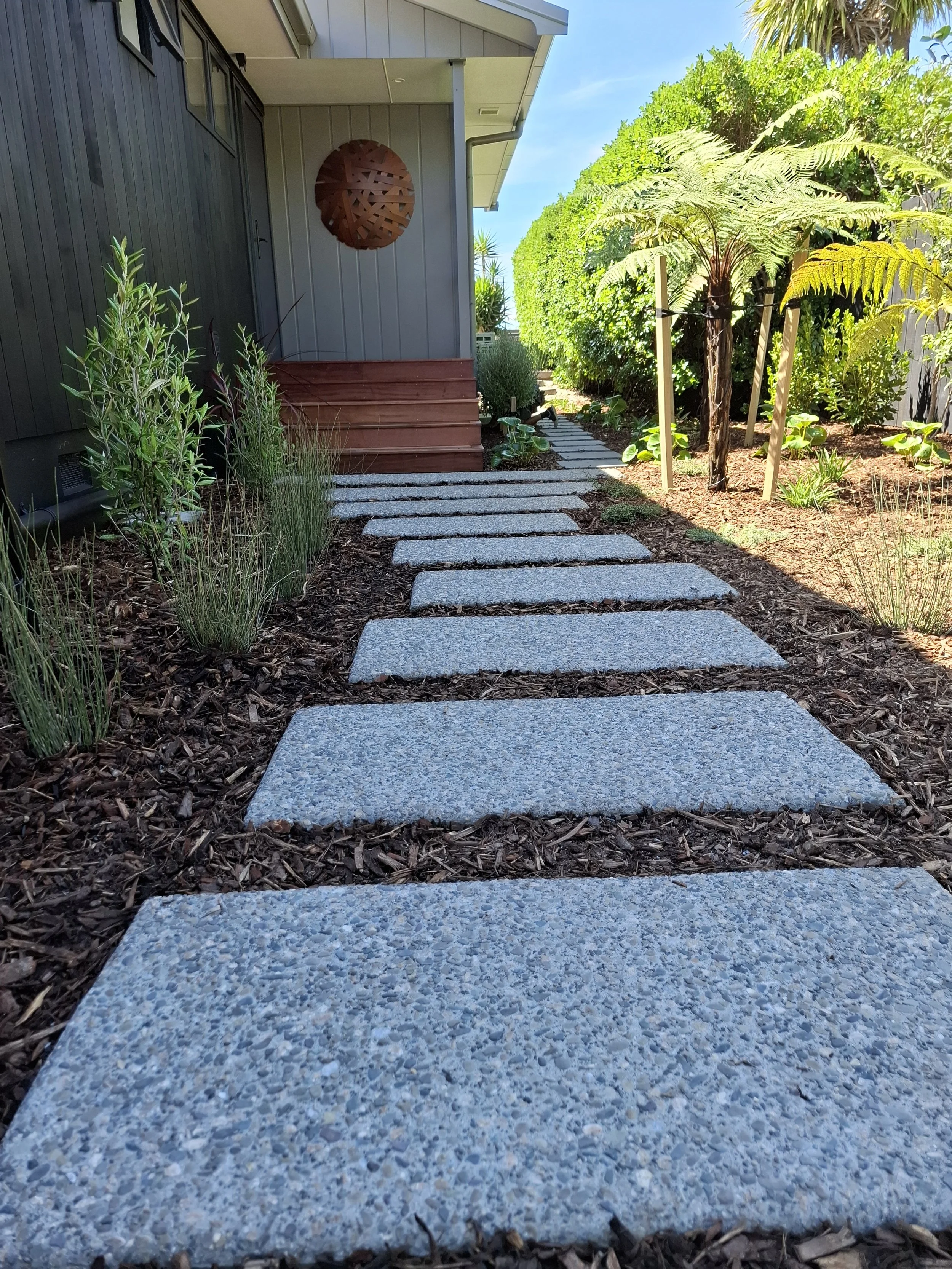 A stone pathway leading to a house with a small set of stairs, surrounded by plants and greenery, in sunny weather.