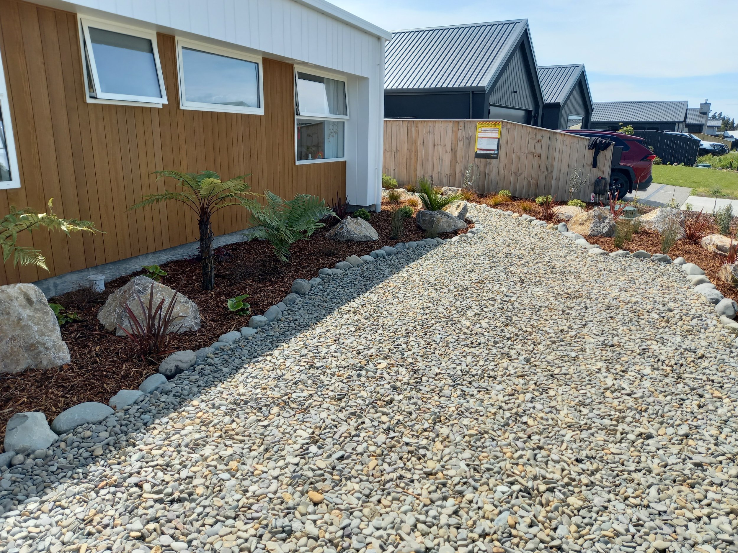 A gravel walkway with large rocks and small plants on the side, leading past a wooden fence and modern houses with metal roofs, parked cars, and landscaped yards.