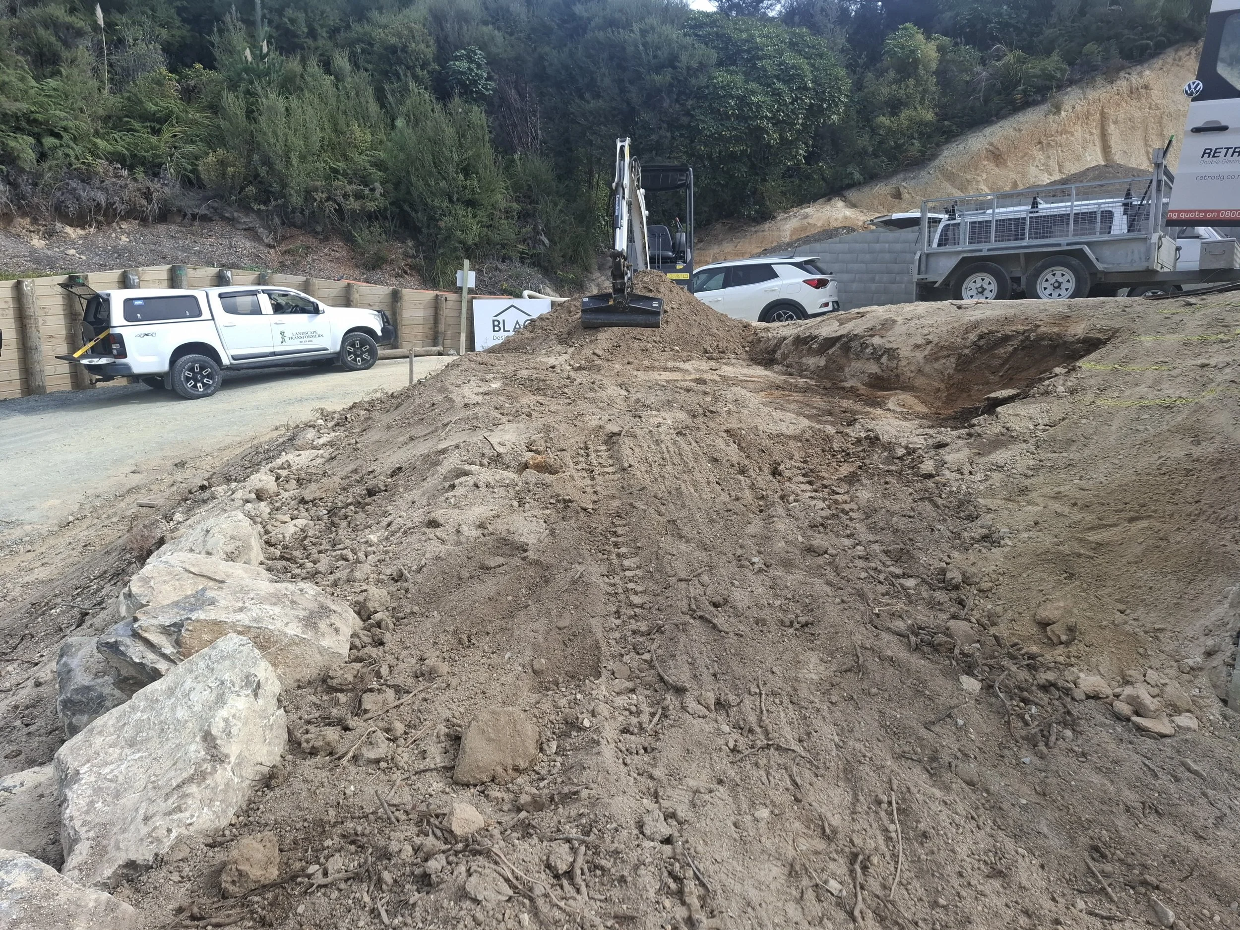 Construction site with excavator digging into dirt and rocks, white cars parked nearby and construction trucks in the background, surrounded by green trees and hillside.