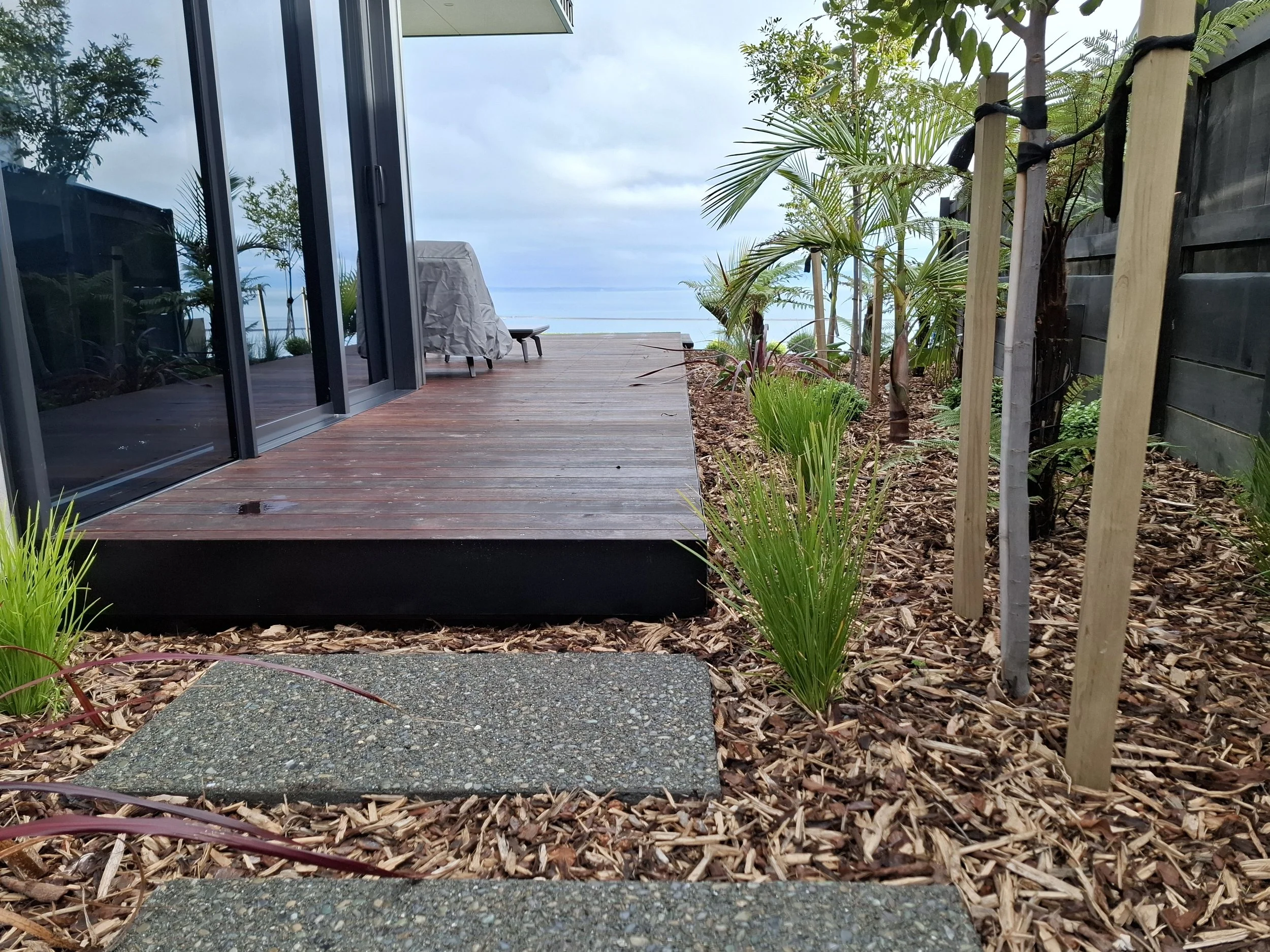 A wooden deck outside a modern house with black metal frame sliding glass doors, surrounded by plants and mulch, overlooking the ocean under cloudy skies.