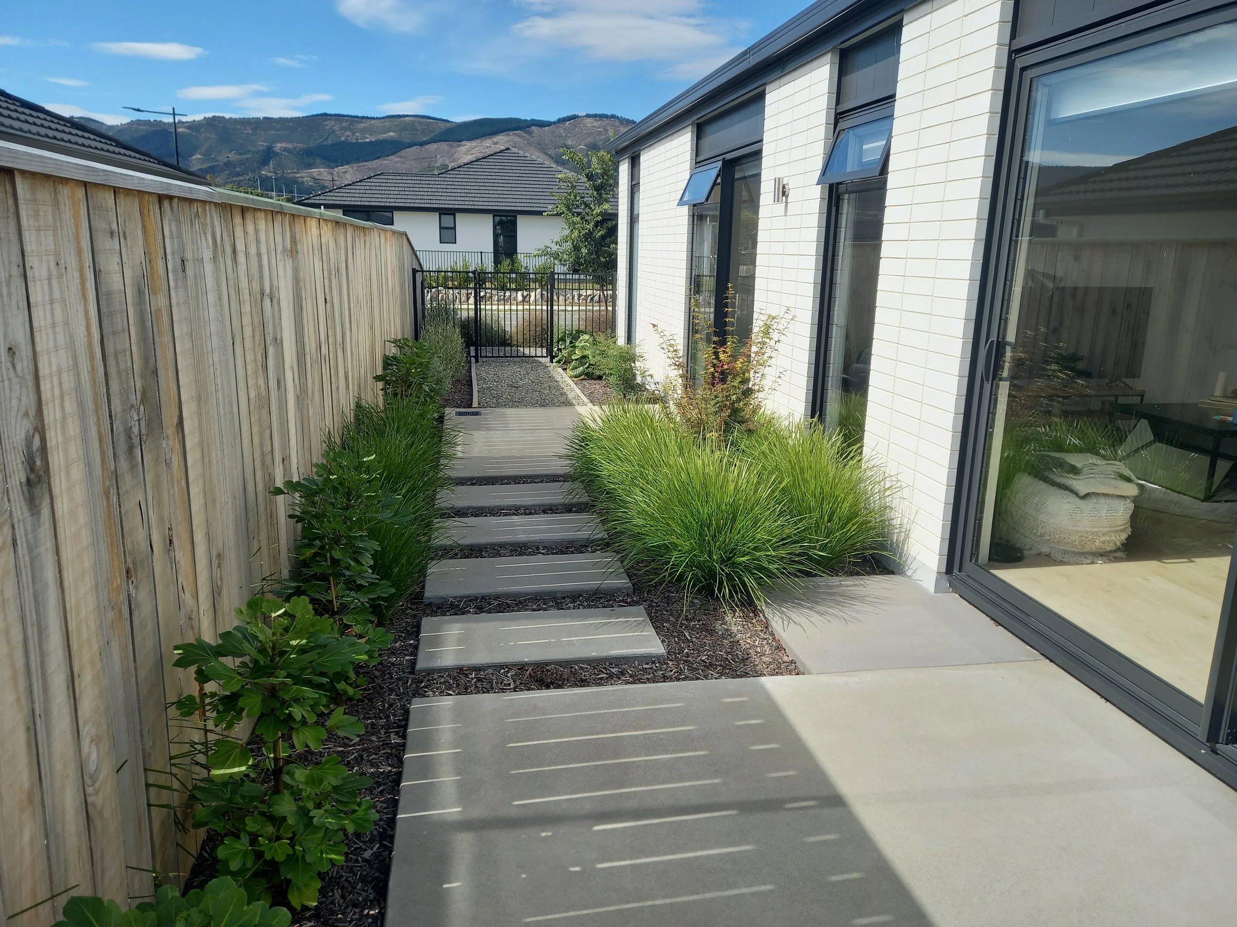 A narrow backyard walkway with concrete stepping stones and a wood border, bordered by lush green plants on one side and a modern white brick house with large windows on the other, with a mountain range in the background under a partly cloudy sky.