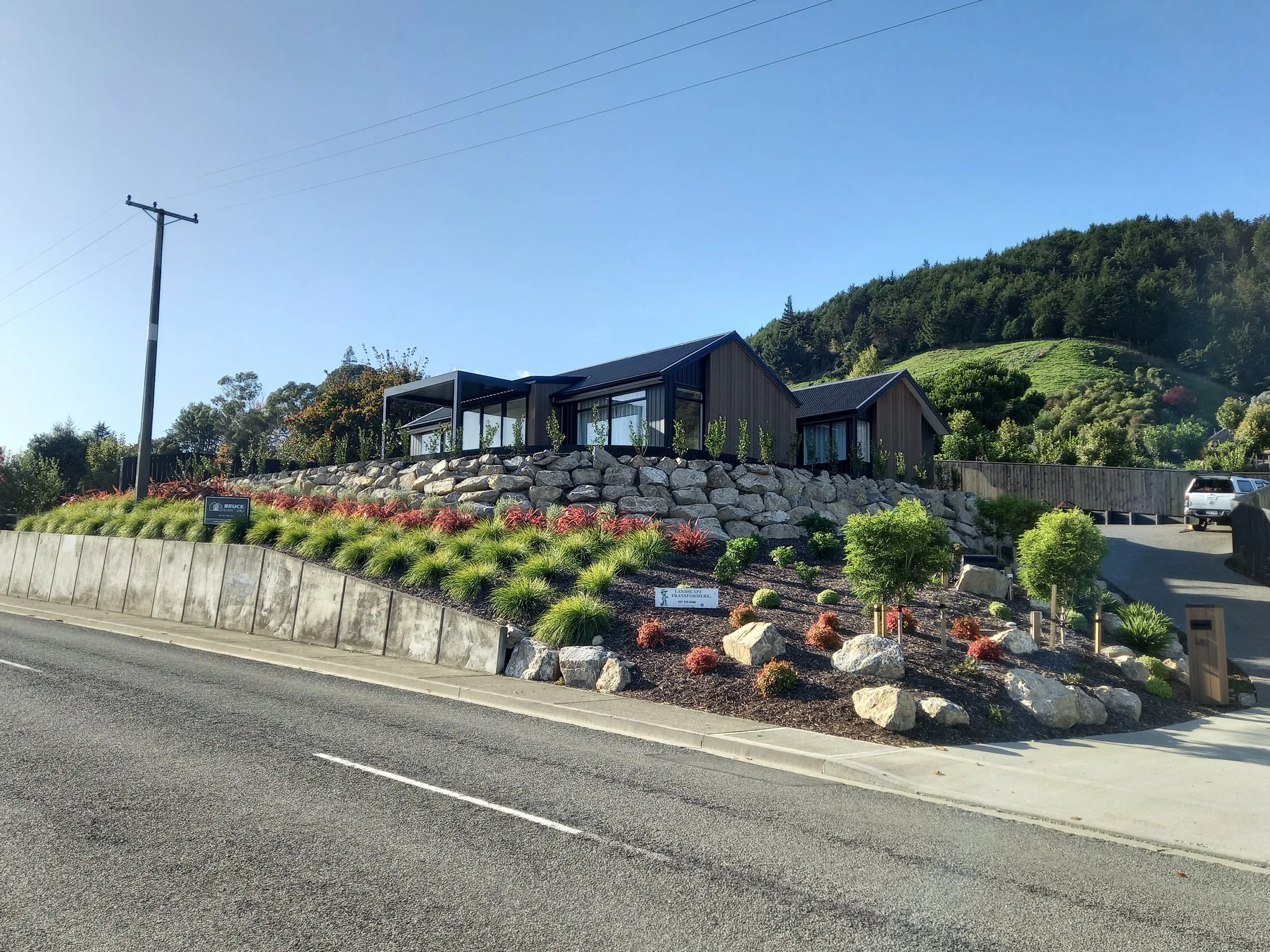 A modern house with large windows and a black roof, set on a hillside with lush greenery and trees. A stone retaining wall and landscaped garden with red and green bushes are in the foreground, with a clear blue sky overhead.