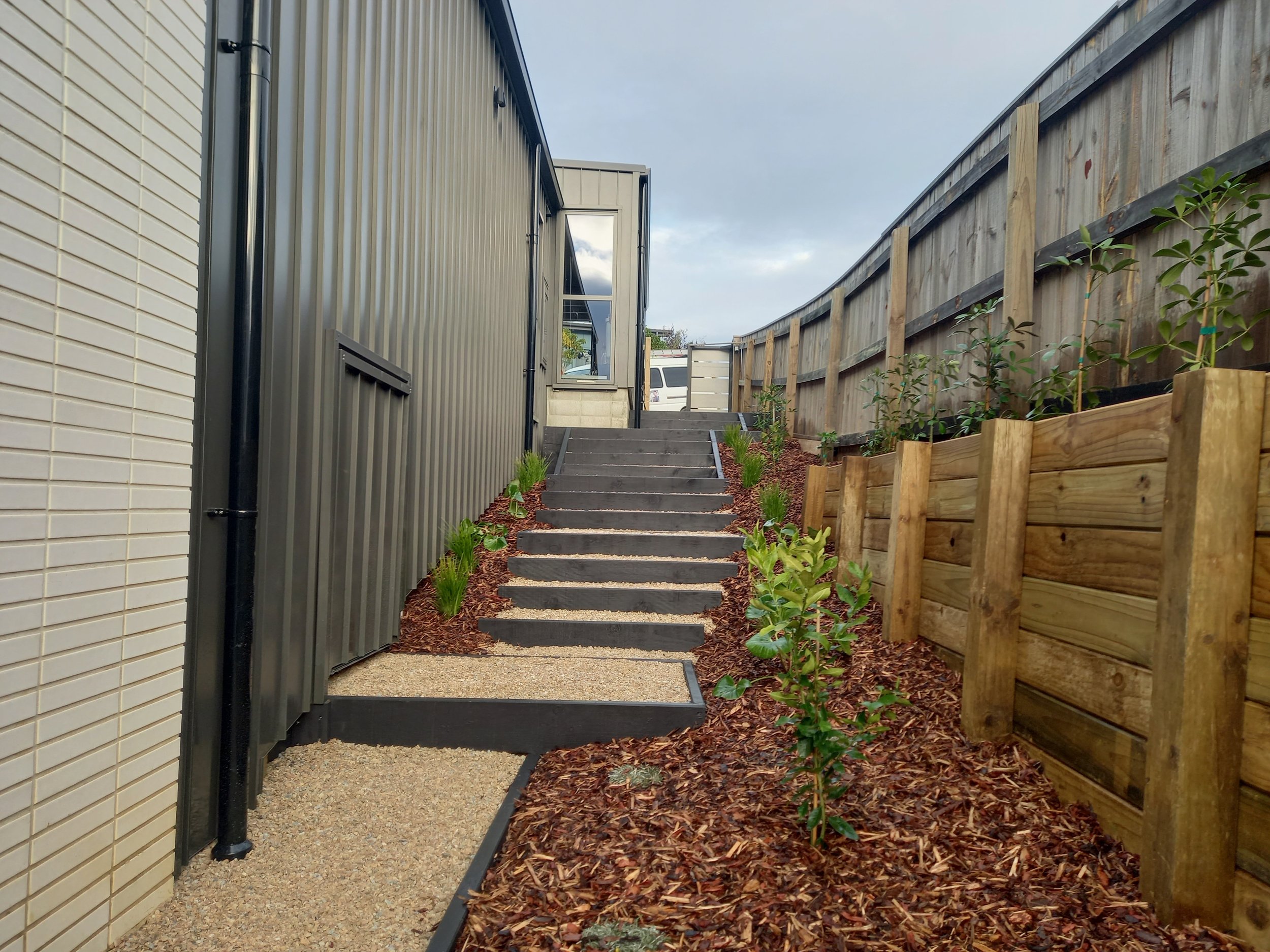 Outdoor staircase leading up between two fenced areas, with plants along the right side and a house wall on the left, under a cloudy sky.