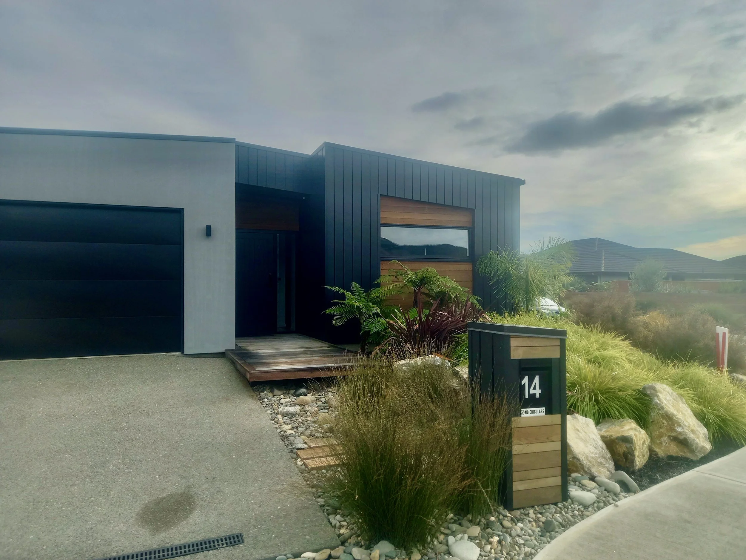Modern house with black, gray, and wood exterior, small front garden with plants, rocks, and a walkway, numbered 14, in a suburban neighborhood under cloudy sky.