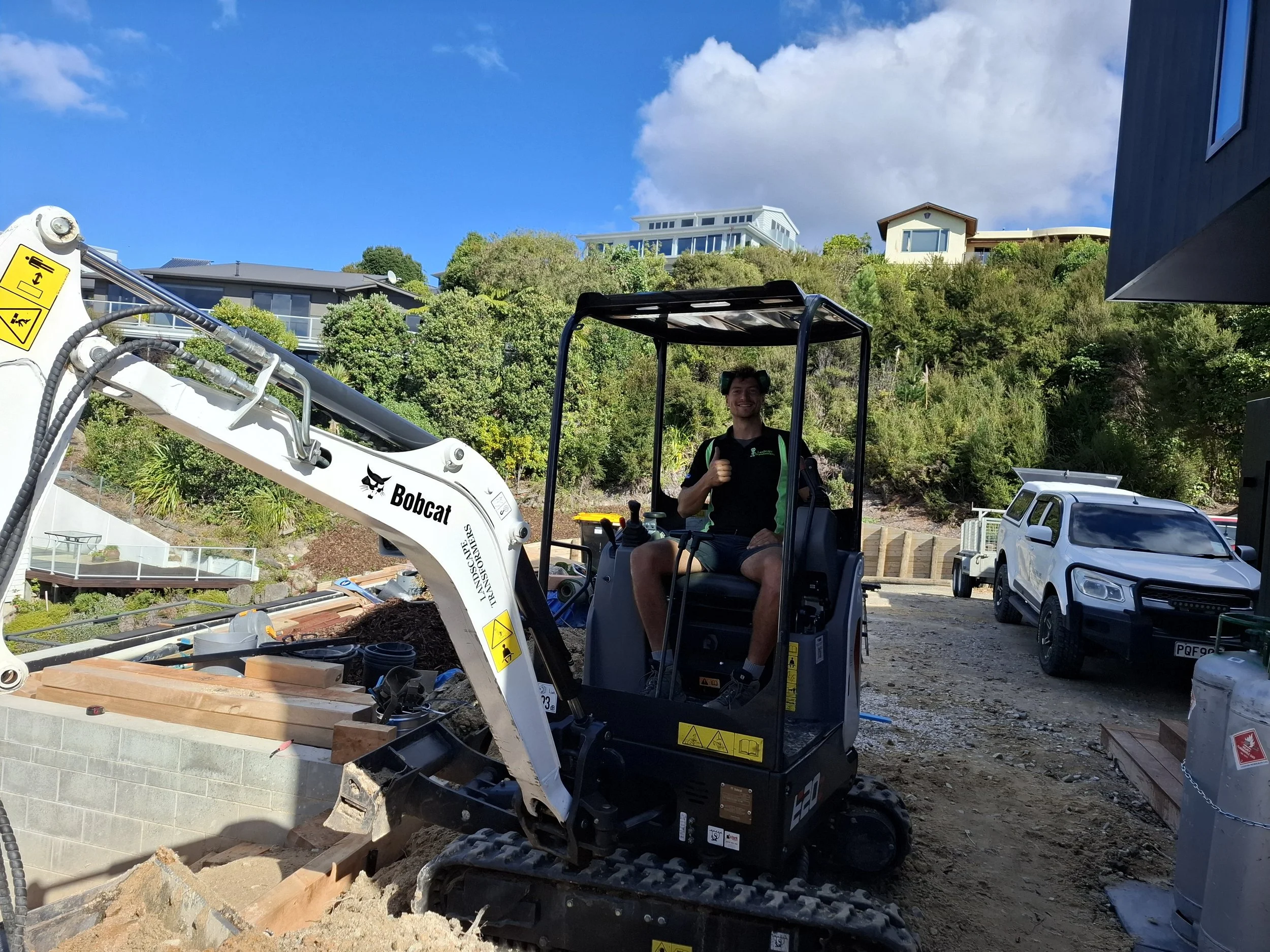 A person sitting on a small construction vehicle, giving a thumbs up. The construction vehicle has a digging arm with a bucket and is labeled 'Bobcat.' There are houses and greenery on a hillside in the background, with a blue sky and clouds overhead