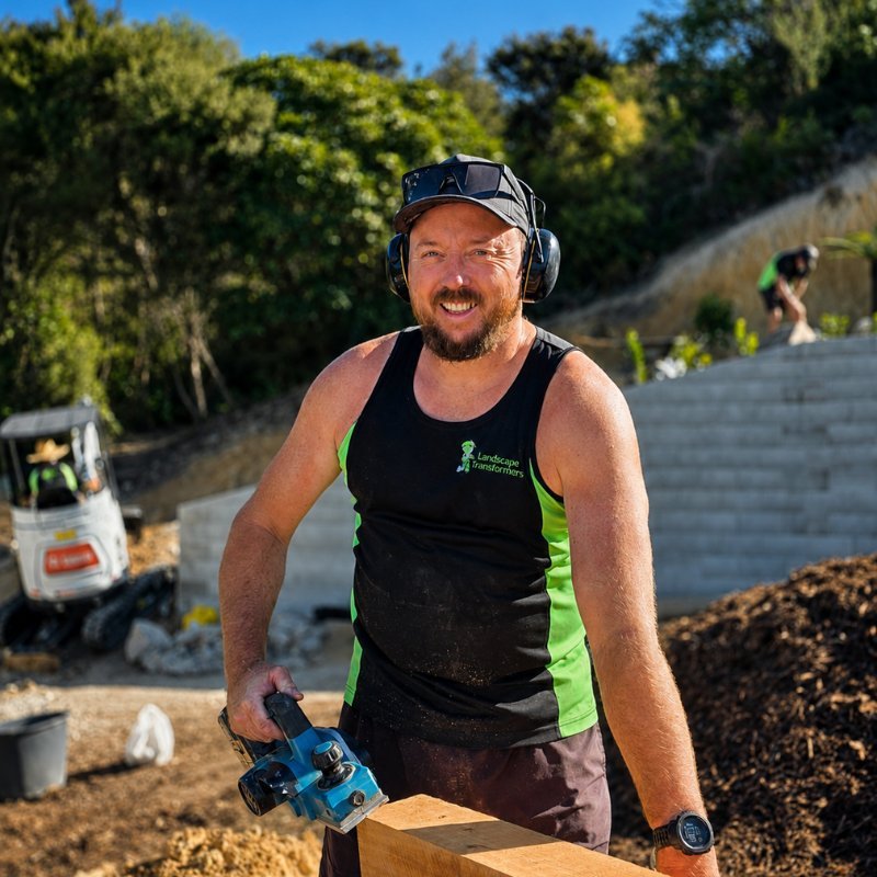 A man working outdoors with safety gear on, holding a cordless power saw, smiling, at a construction site with construction equipment and workers in the background.