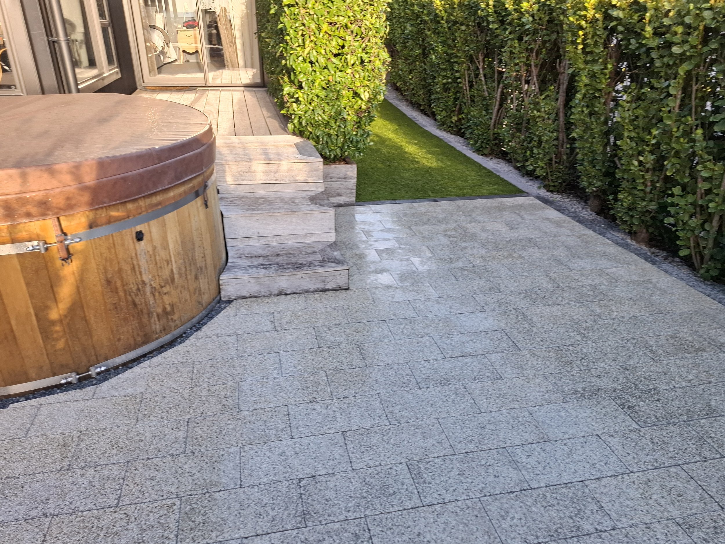 A patio area with stone flooring, a wooden hot tub, and a sliding glass door leading into a house. There is a green hedge and small grass patch along one side.