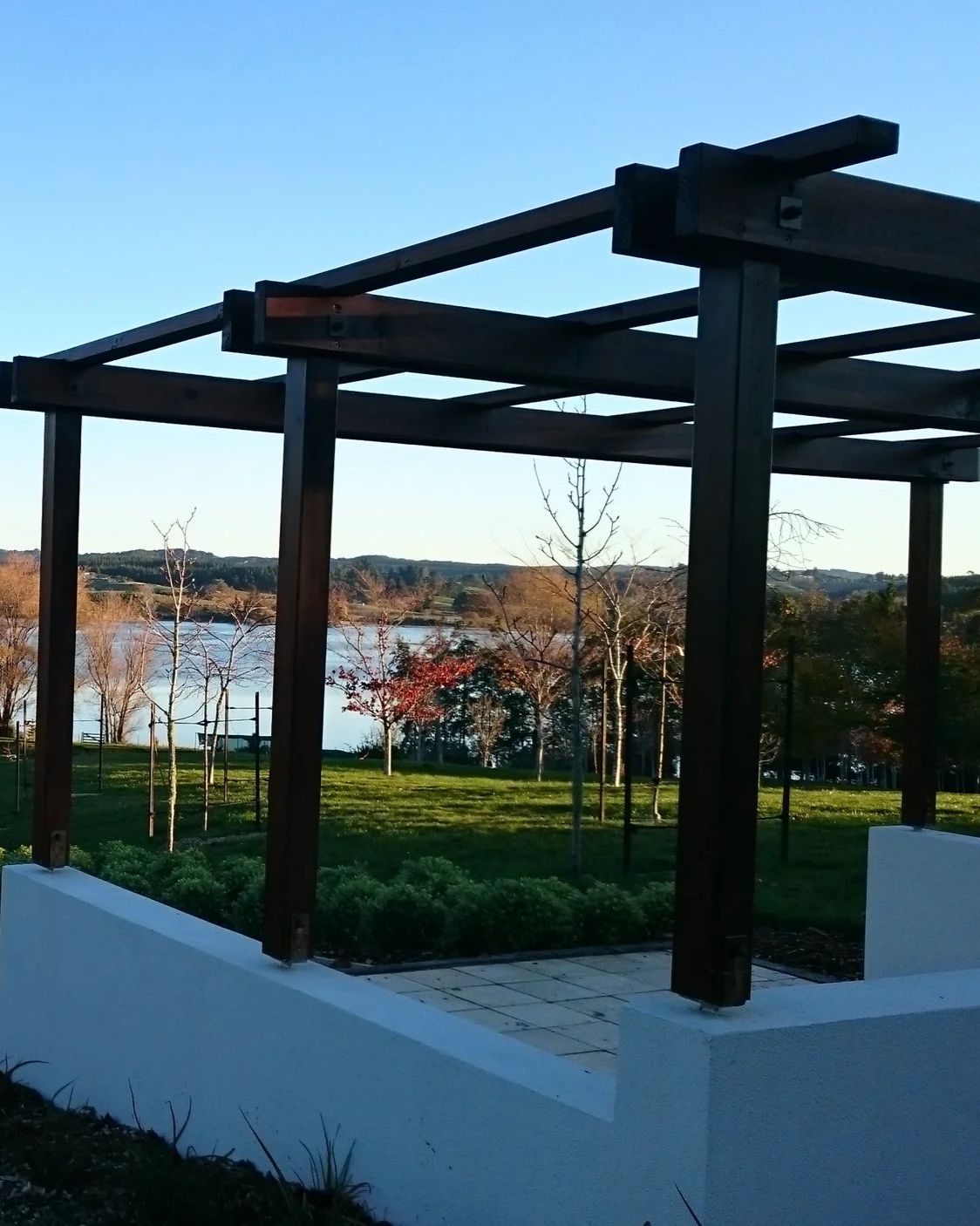 Wooden pergola overlooking a park with trees, grass, and a river in the background during late afternoon.