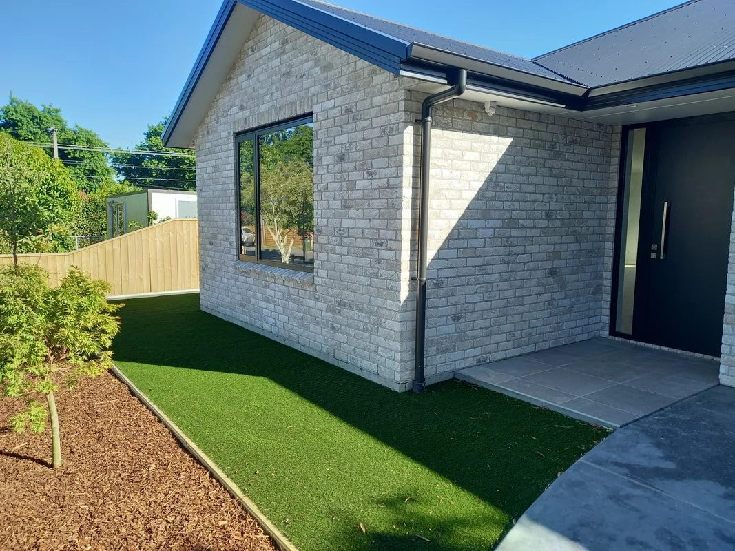 Exterior of a modern house with light gray brick walls, large window, black door with handle, covered porch, synthetic grass lawn, wooden fence, and trees in the background under a blue sky.