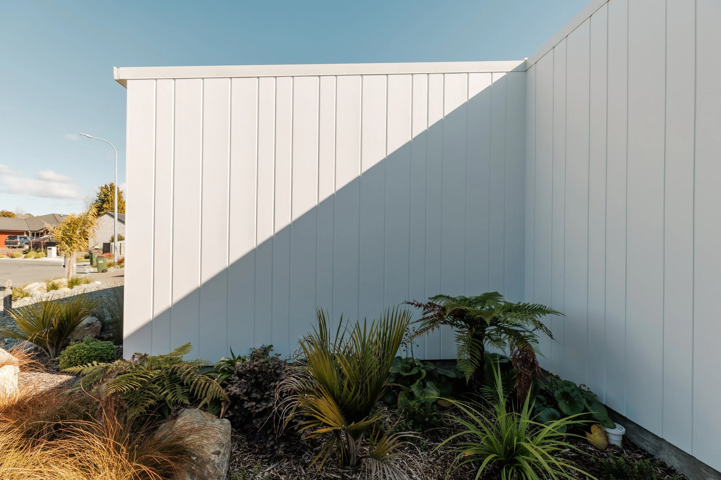 White building wall with a shadow cast across it, surrounded by landscaped plants and rocks in a residential neighborhood.