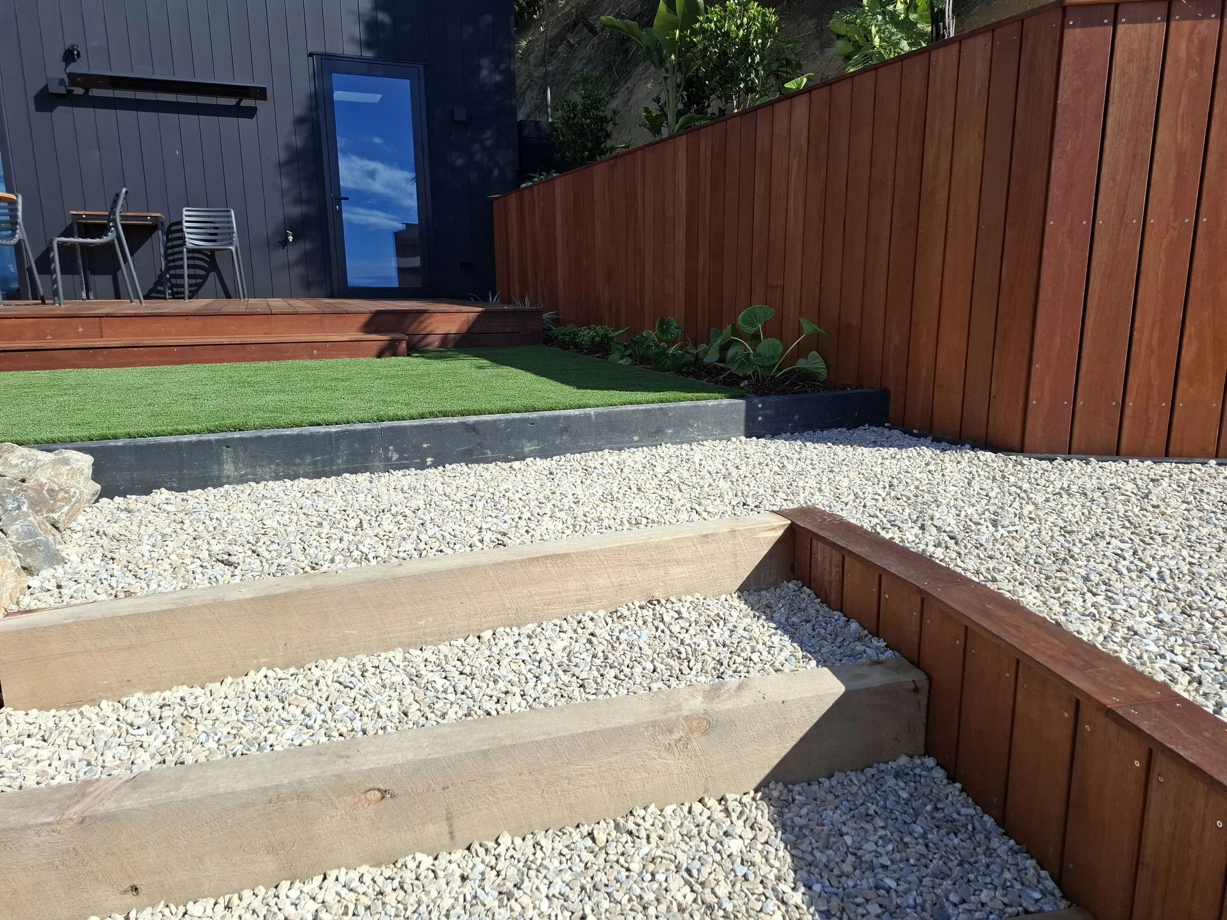 A backyard patio with wooden stairs leading to a grassy area, surrounded by a red wooden fence, with some plants along the fence line. There are chairs and a table near a dark-colored building with a door and a window reflecting the blue sky.