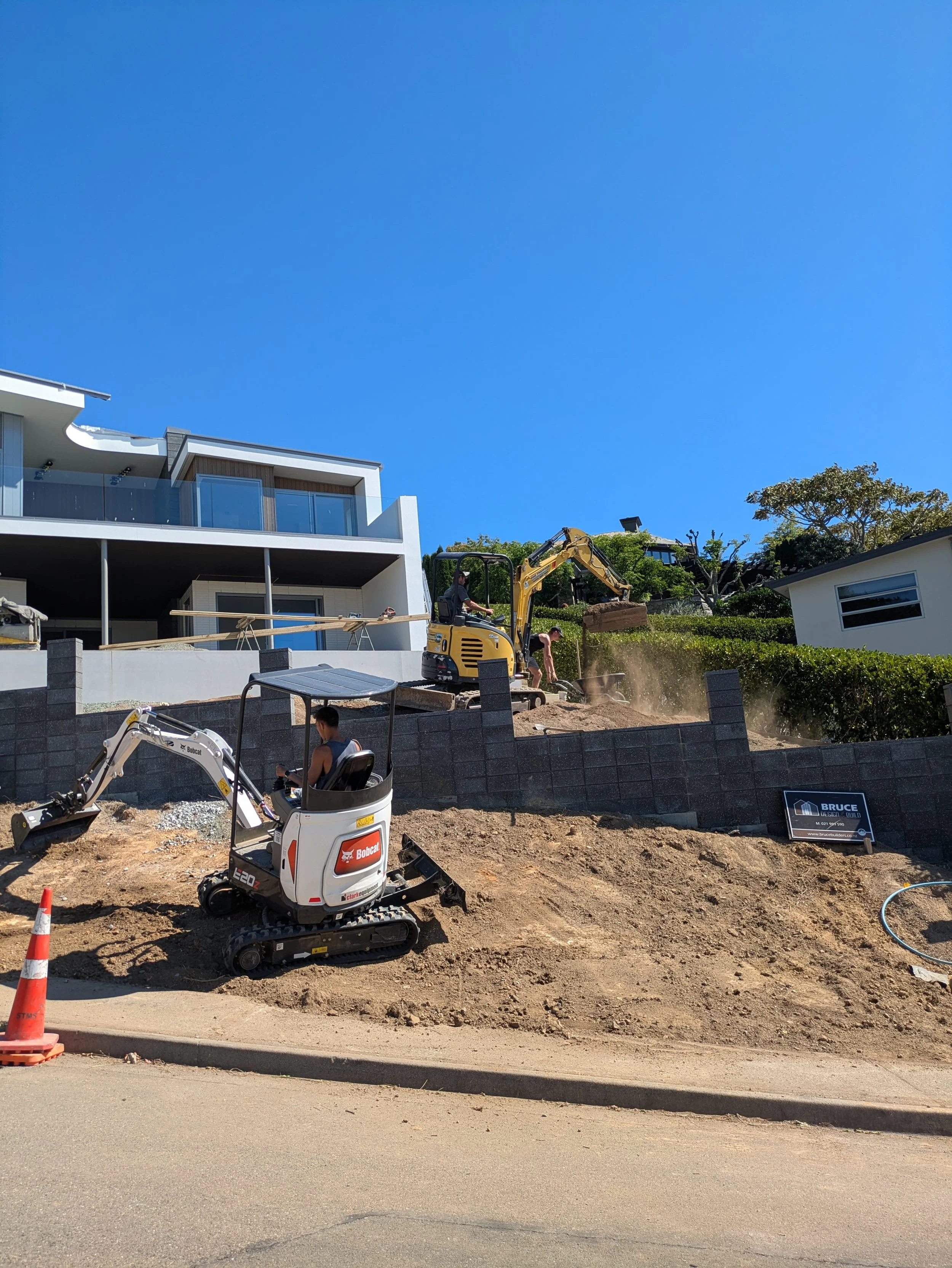 Construction site in a residential area with workers operating small machinery on a dirt hill, modern house with large glass windows and balconies on the left, trees on the right, clear blue sky overhead, and orange traffic cones at the edge of the r