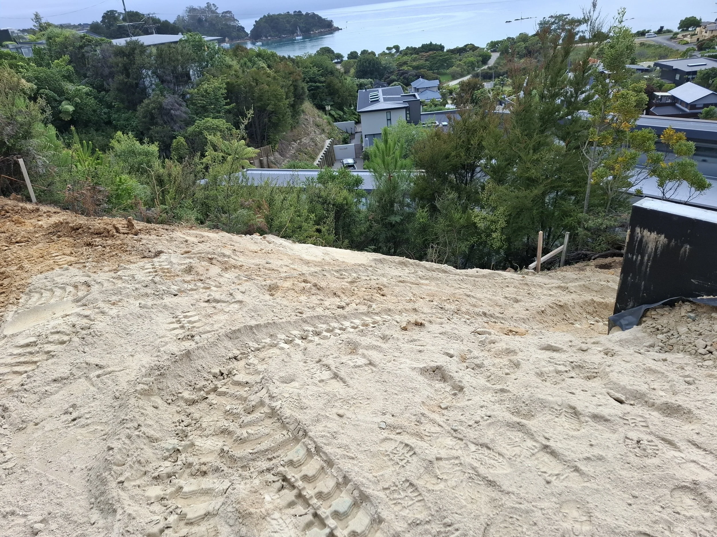 A dirt construction site with visible tire tracks in the foreground, overlooking a lush green residential area near a body of water with boats and trees in the background.