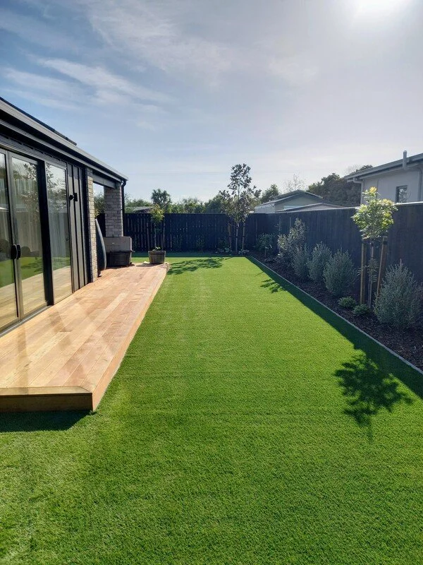 Backyard with a wooden patio, glass sliding doors, green grass lawn, potted plants, and trees along the fence under a partly cloudy sky.