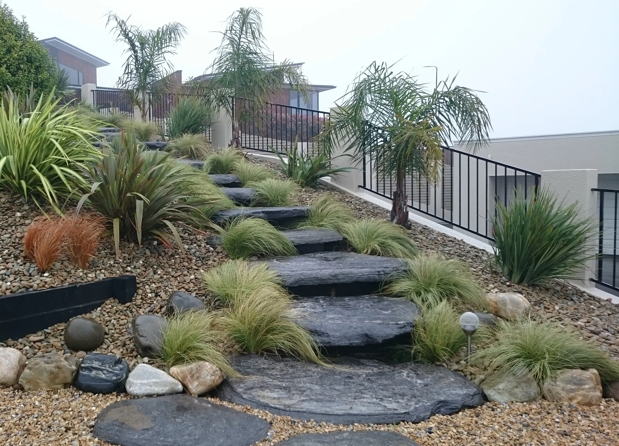 A landscaped garden with black stone steps and a water feature, surrounded by various plants including grasses and tall palm-like trees, with modern houses in the background and a foggy sky.