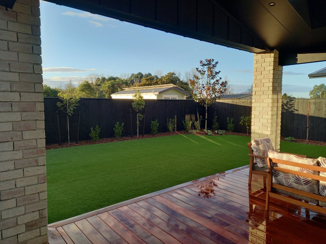 View of a backyard with a wooden deck, green grass lawn, young trees, and a black fence against a blue sky with some clouds.