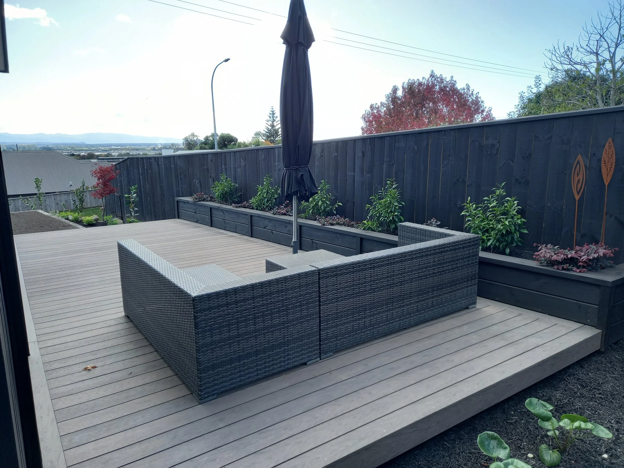 A backyard deck with outdoor wicker furniture and a closed patio umbrella, bordered by a black wooden fence with decorative designs, overlooking a view of the neighborhood and mountains in the distance.