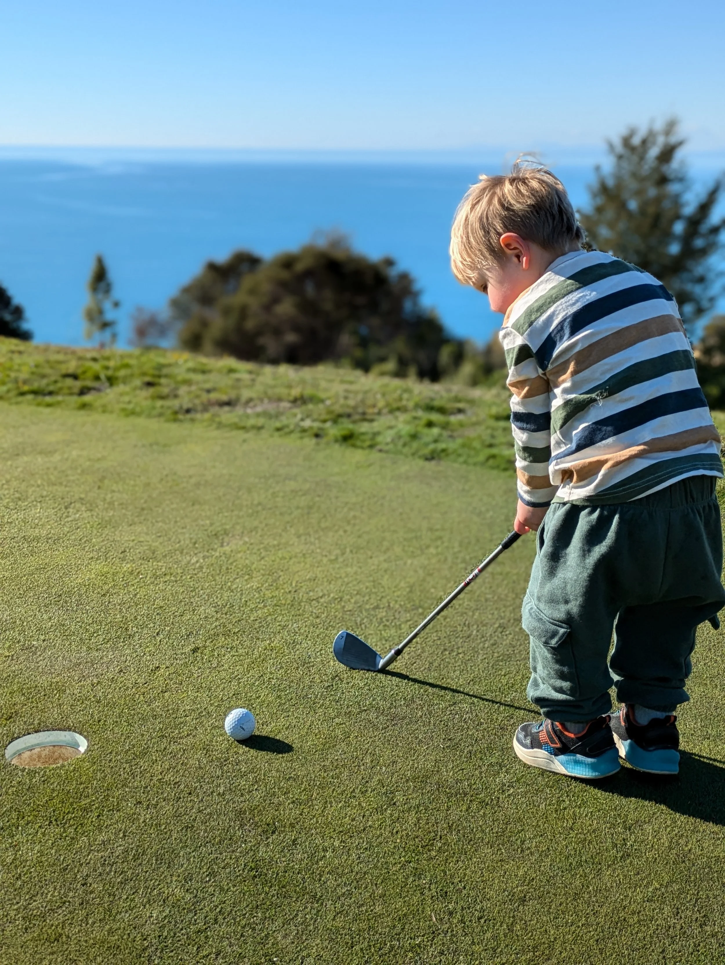 A young child preparing to putt a golf ball on a golf course, with a hole and a golf ball visible on the green grass, and trees and blue sky in the background.