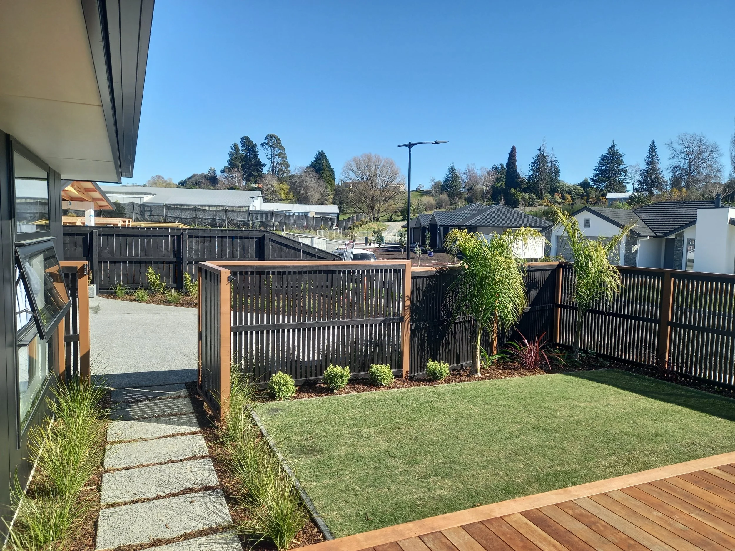 View of a backyard patio area with a small grassy patch, a wooden deck, and a stone pathway. There is a black wooden fence, a few plants, a hill with trees, and neighboring houses visible in the background under a clear blue sky.