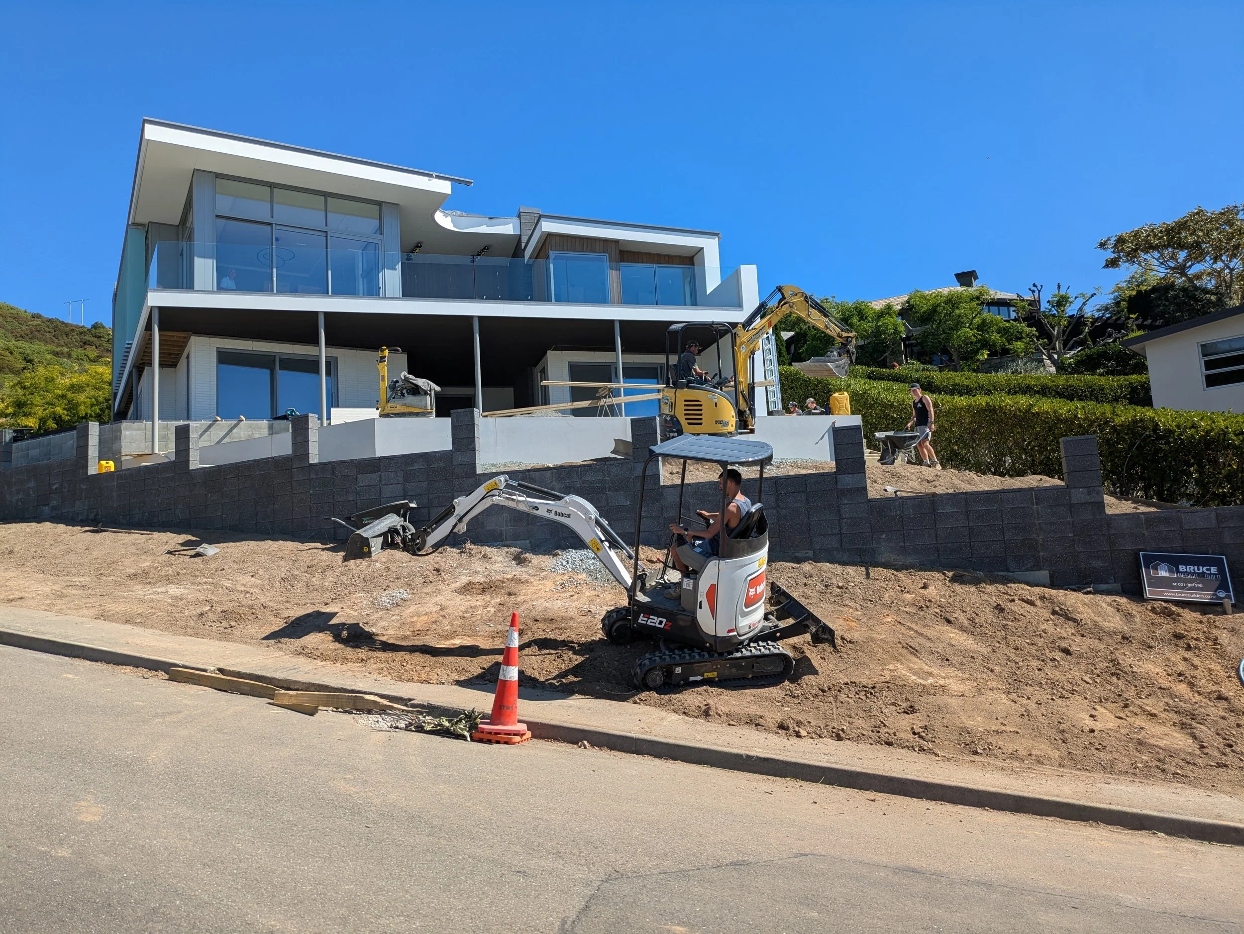 Construction site with a modern house under construction, a small excavator, workers, a temporary fence, a traffic cone, and a clear blue sky.