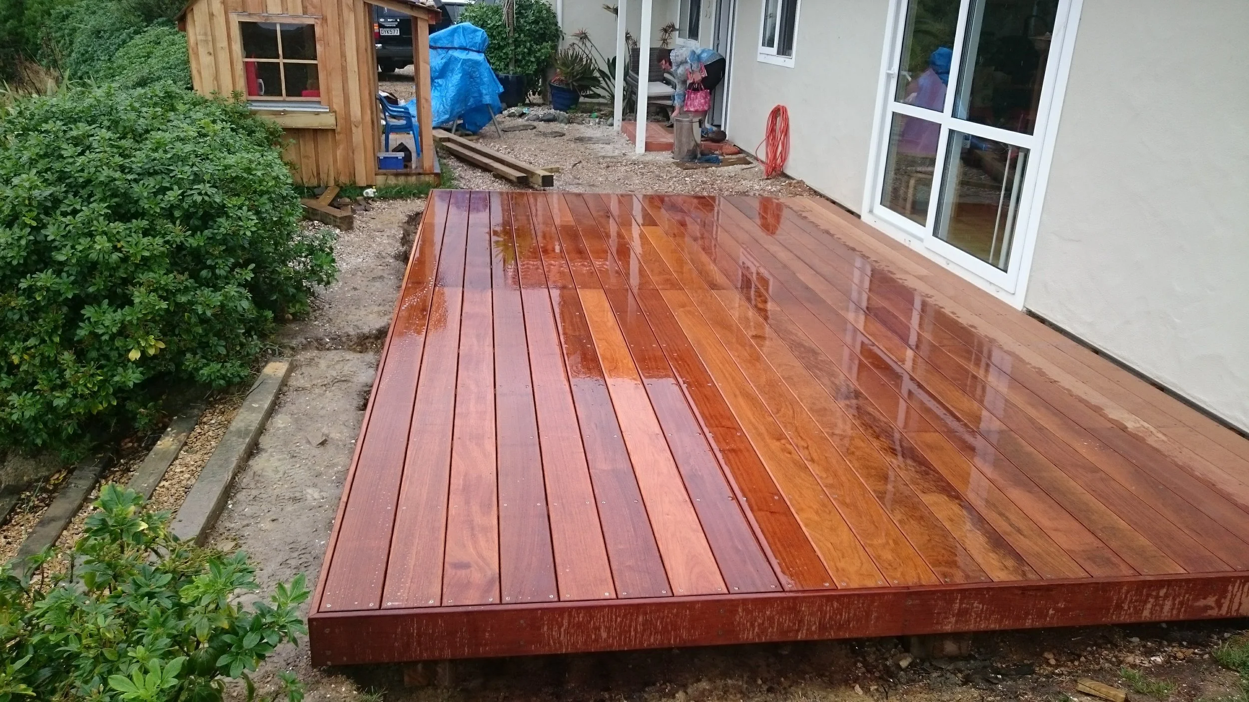 A newly built wooden deck outside a house with wet surface from rain, reflecting parts of surrounding objects, with a garden area and an unfinished section nearby.
