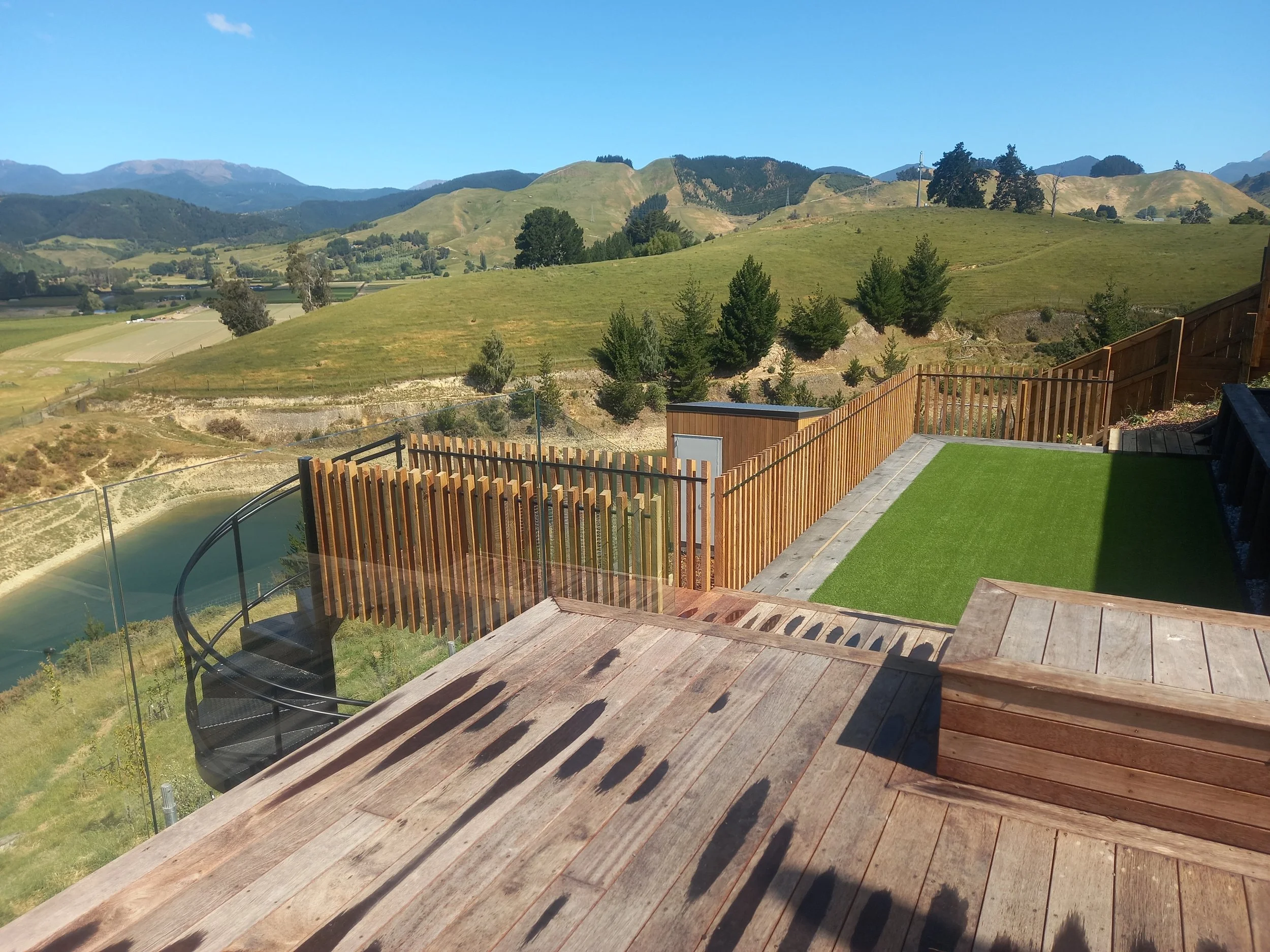 View from a house balcony overlooking rolling green hills with scattered trees, mountains in the distance under a clear blue sky.