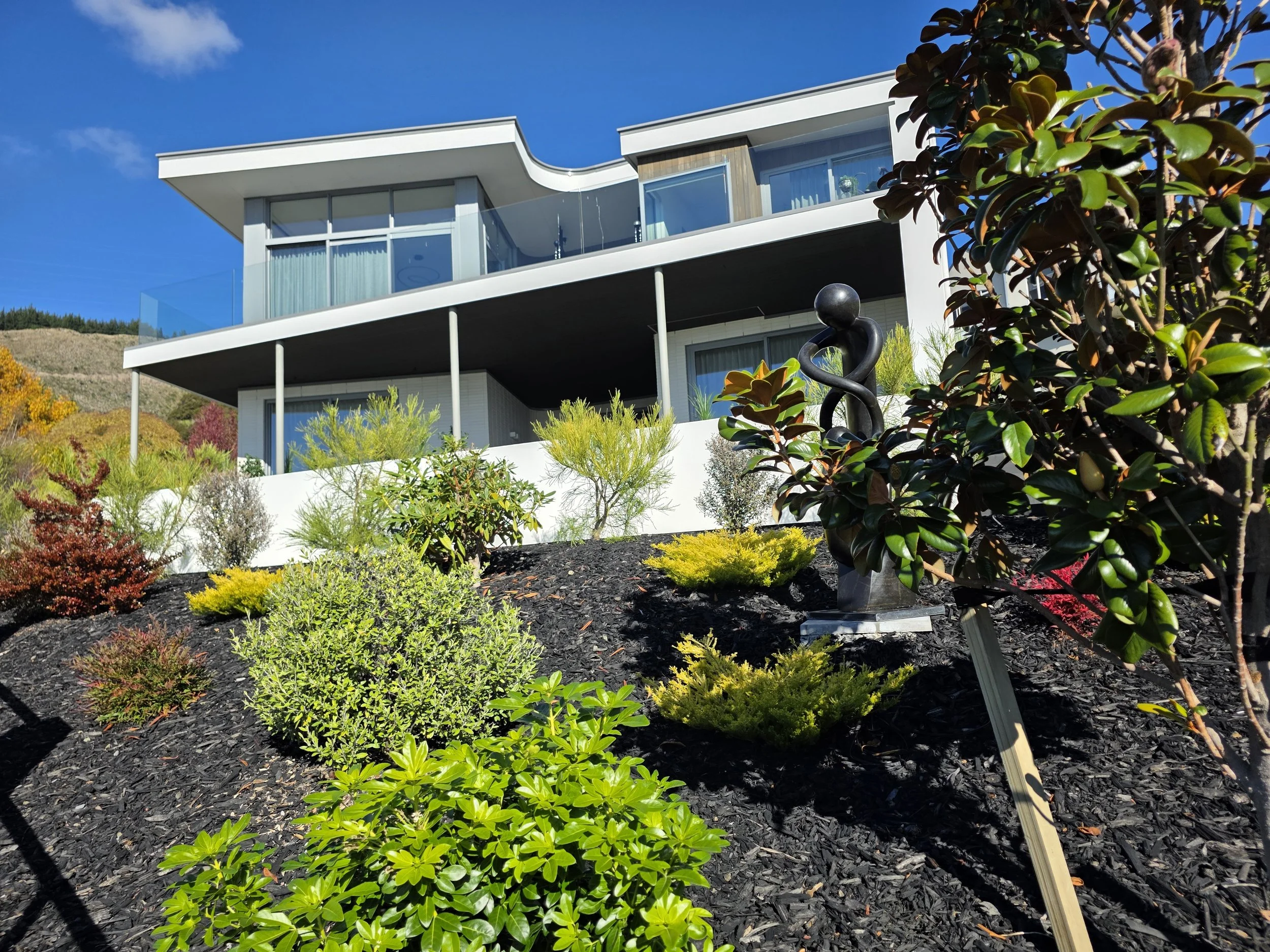 Modern house with a large balcony, glass railing, and large windows, surrounded by lush landscaping and colorful plants under a clear blue sky.
