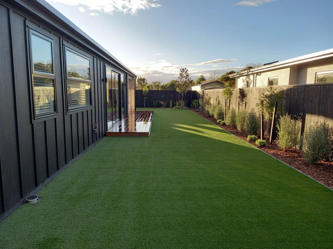 Backyard view with artificial grass, small deck, and garden bed along the fence