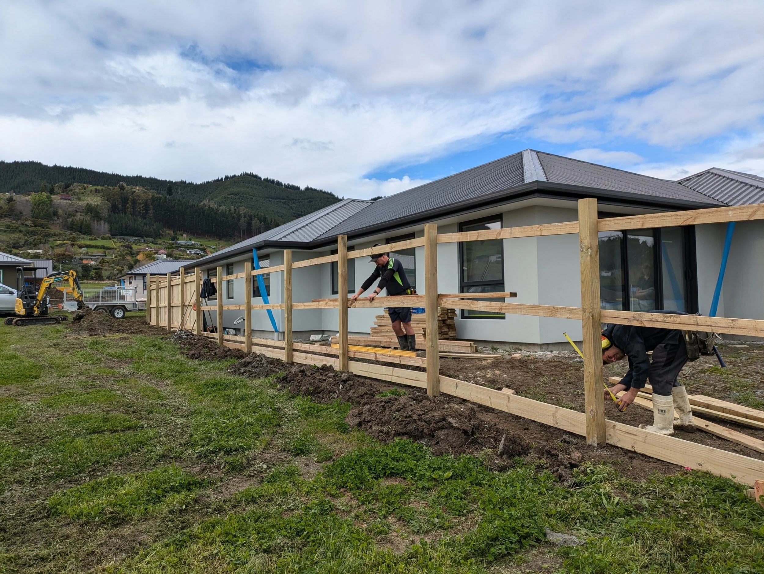 Construction workers building a wooden fence in front of a modern house with a metal roof, with green hills and a partly cloudy sky in the background.