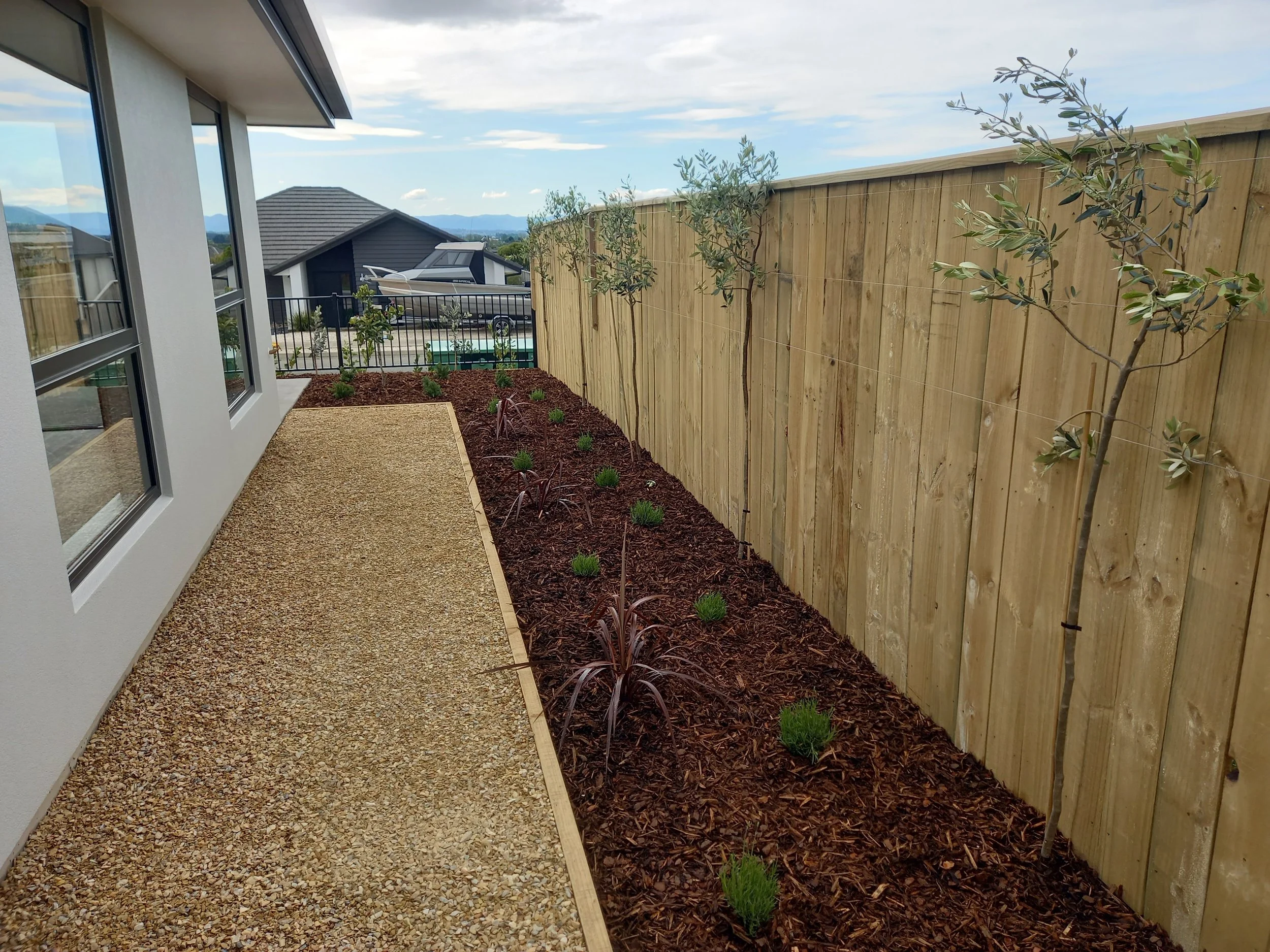 View of a backyard with a gravel patio edge, young trees along a wooden privacy fence, neighboring houses, and distant mountains under a partly cloudy sky.