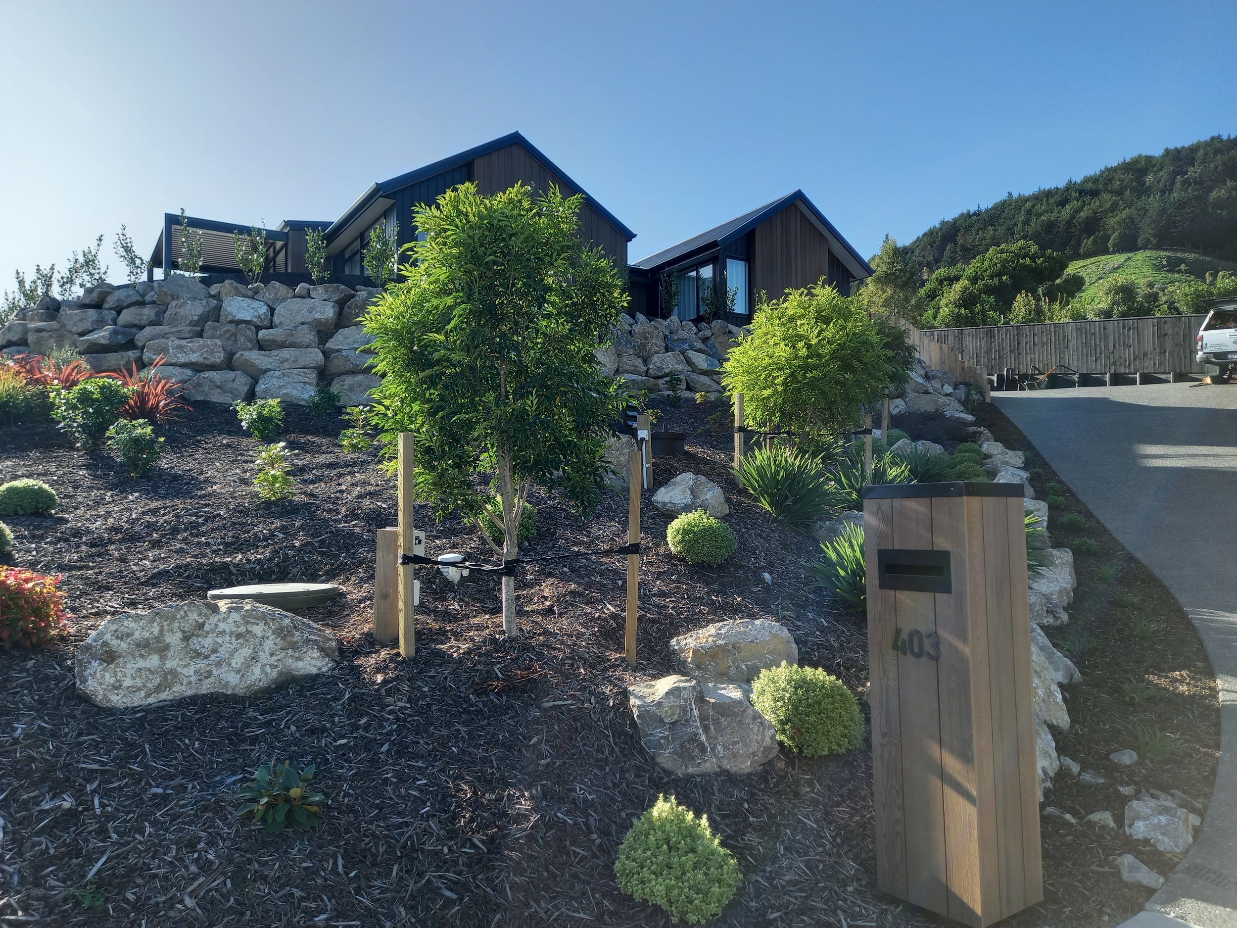 Modern house on a hill with rocks and lush landscaped garden, driveway, and car.