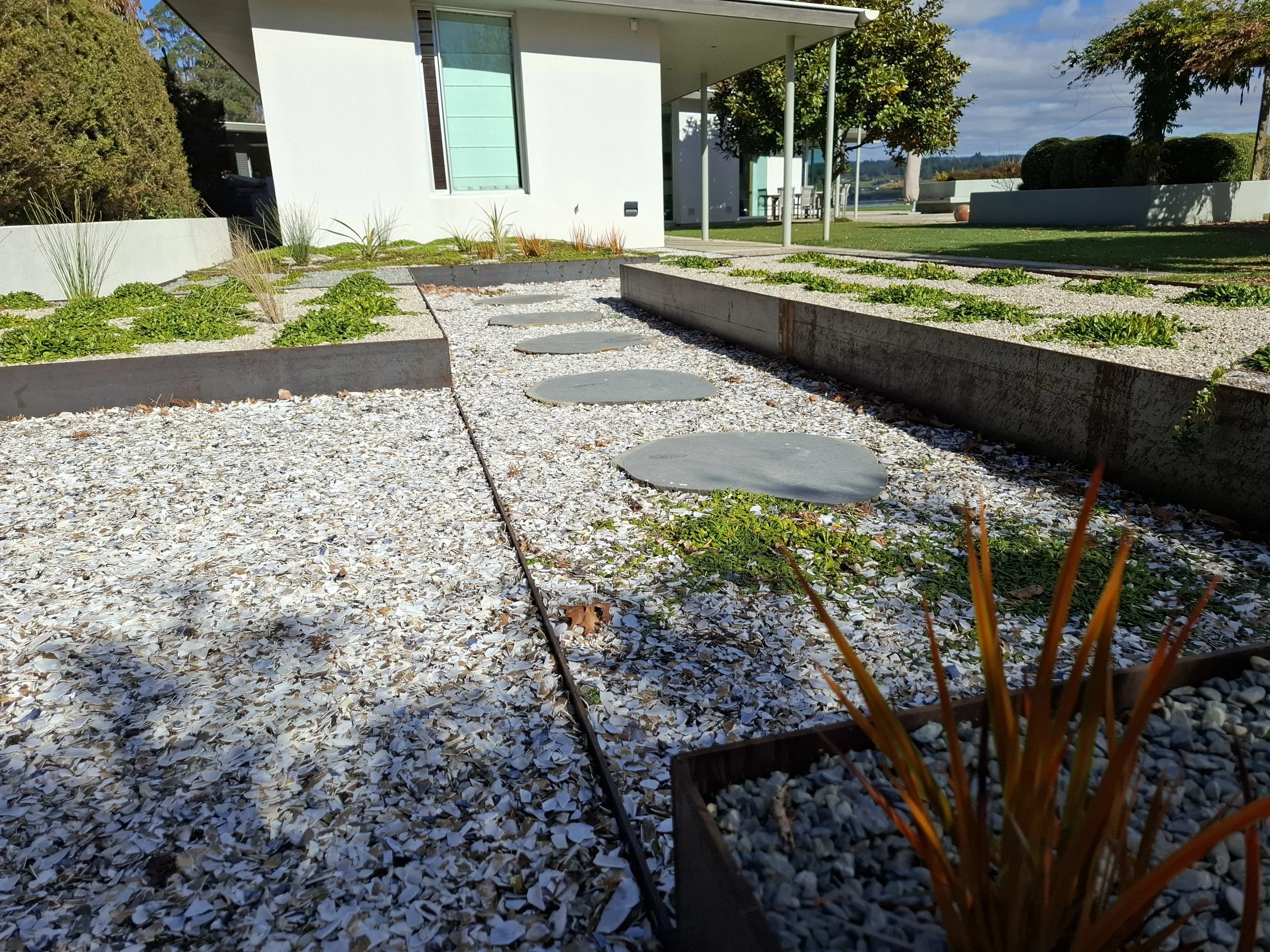 Modern residential house exterior with a small garden path, gravel ground cover, and landscaped bushes and trees under a partly cloudy sky.