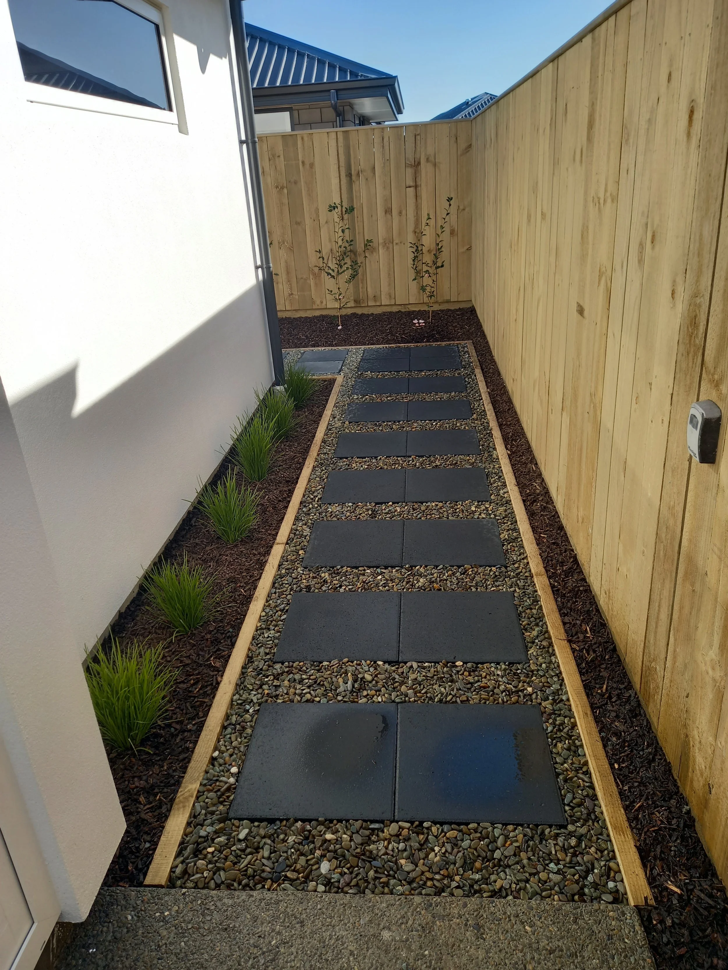 A small outdoor walkway with rectangular stepping stones set in gravel, bordered by a wooden frame, next to a house with a white exterior wall and a window. On the right side, there is a light wooden privacy fence with two young trees planted in fron