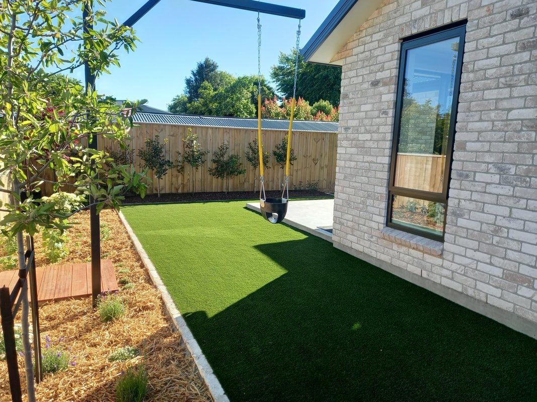 A backyard with a swing set, artificial grass, a brick house, a wooden fence, some trees and plants, on a sunny day.