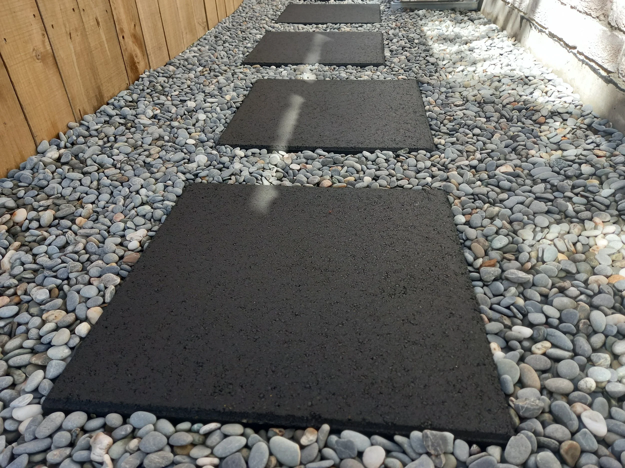 A pathway with square black stepping stones surrounded by small multicolored pebbles, running alongside a wooden fence on the left and a concrete wall on the right.