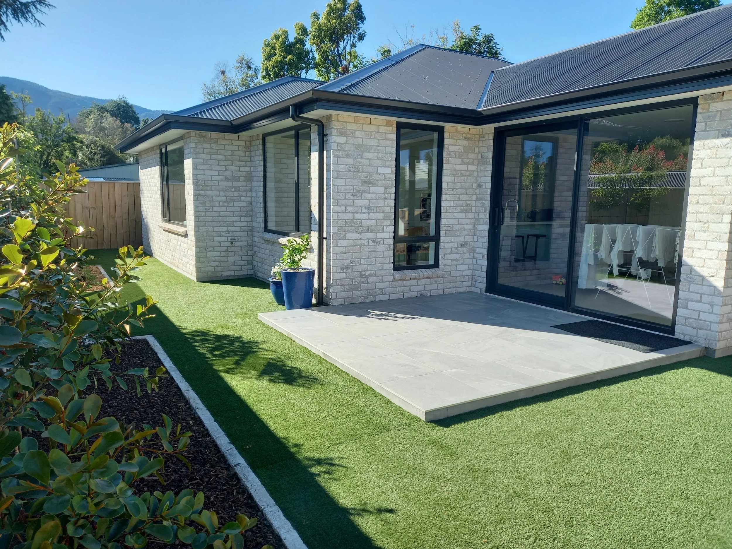 A modern house with white brick walls and large glass doors and windows. There is a backyard with a small lawn, potted plants, and a paved patio area. The house has a dark metal roof, and trees and mountains are visible in the background under a clea