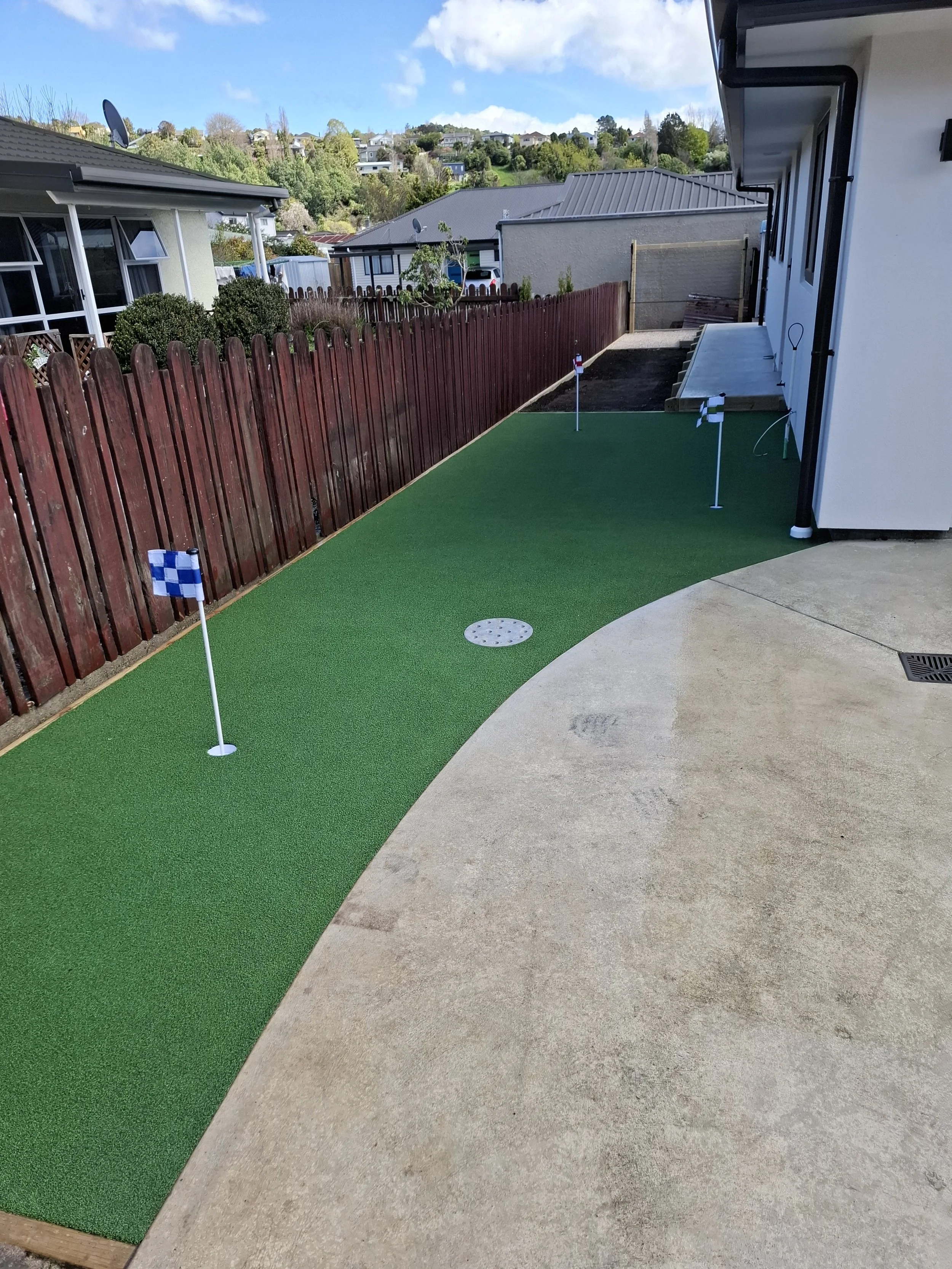 Backyard with putting green, small flags, and a concrete patio, with houses and hills in the background.