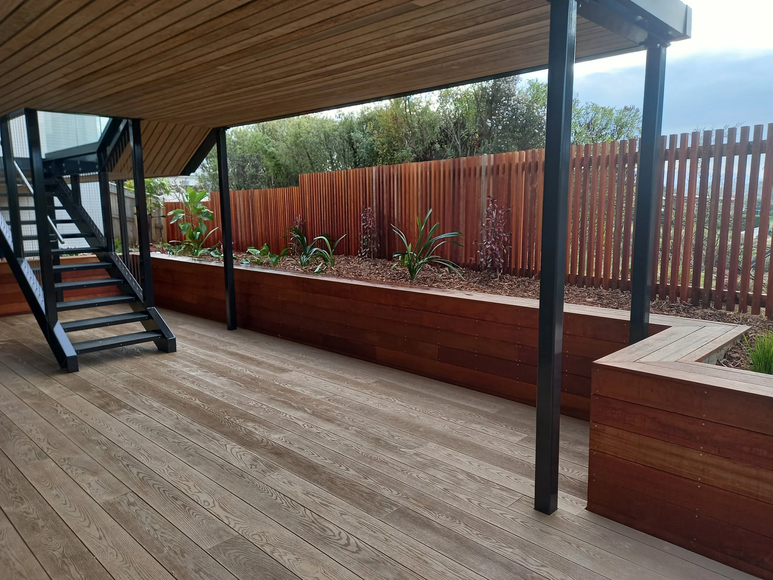 Outdoor balcony with wooden flooring, a raised garden bed with plants, a wooden fence, and a view of trees and sky outside.